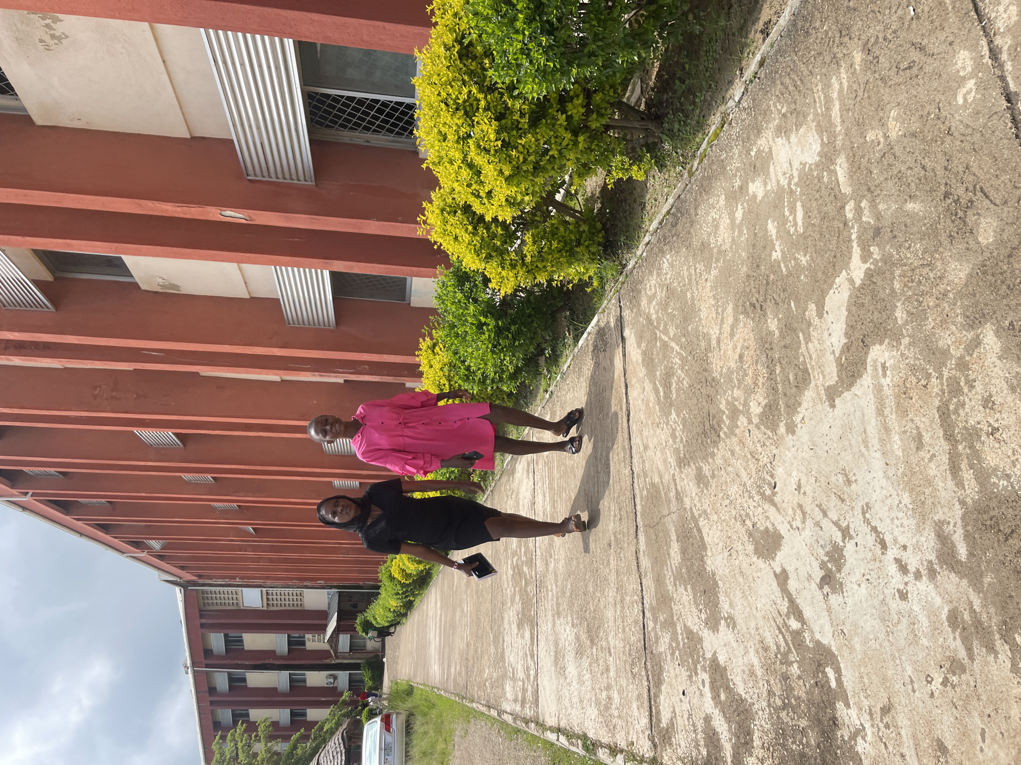 Two individuals are walking on a concrete path adjacent to a large, red, multi-story building. The person on the left is wearing a black dress and a black headscarf, carrying a black item in their right hand. The person on the right is wearing a bright pink dress. Both individuals are walking towards the viewer, and their faces are visible. The building has a distinctive architectural style with vertical red pillars and light-colored sections with windows. In front of the building, there is a row of green and yellow bushes. To the left of the path, there is grass and some trees, and a car is visible in the distance. The sky is overcast with clouds. The scene appears to be outdoors on a sunny day. This is likely a university campus or institutional building in Jos, Nigeria, given the context.