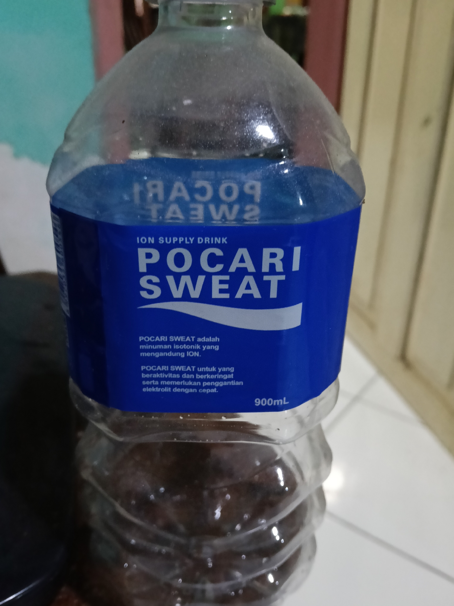 A clear plastic bottle of Pocari Sweat is shown, with a blue label. The bottle is partially filled with a dark liquid. The label reads "ION SUPPLY DRINK" and "POCARI SWEAT" in white text. Below that, there is smaller text in Indonesian that translates to "POCARI SWEAT is an isotonic drink containing ION. POCARI SWEAT for those who are active and sweating and need to replenish ION electrolytes quickly." The volume indicated on the bottle is 900mL. The bottle is standing upright. The background consists of a wall with vertical paneling. The image appears to have been taken indoors, possibly in a home environment. There is no indication of the location being the city of Raha, Indonesia.