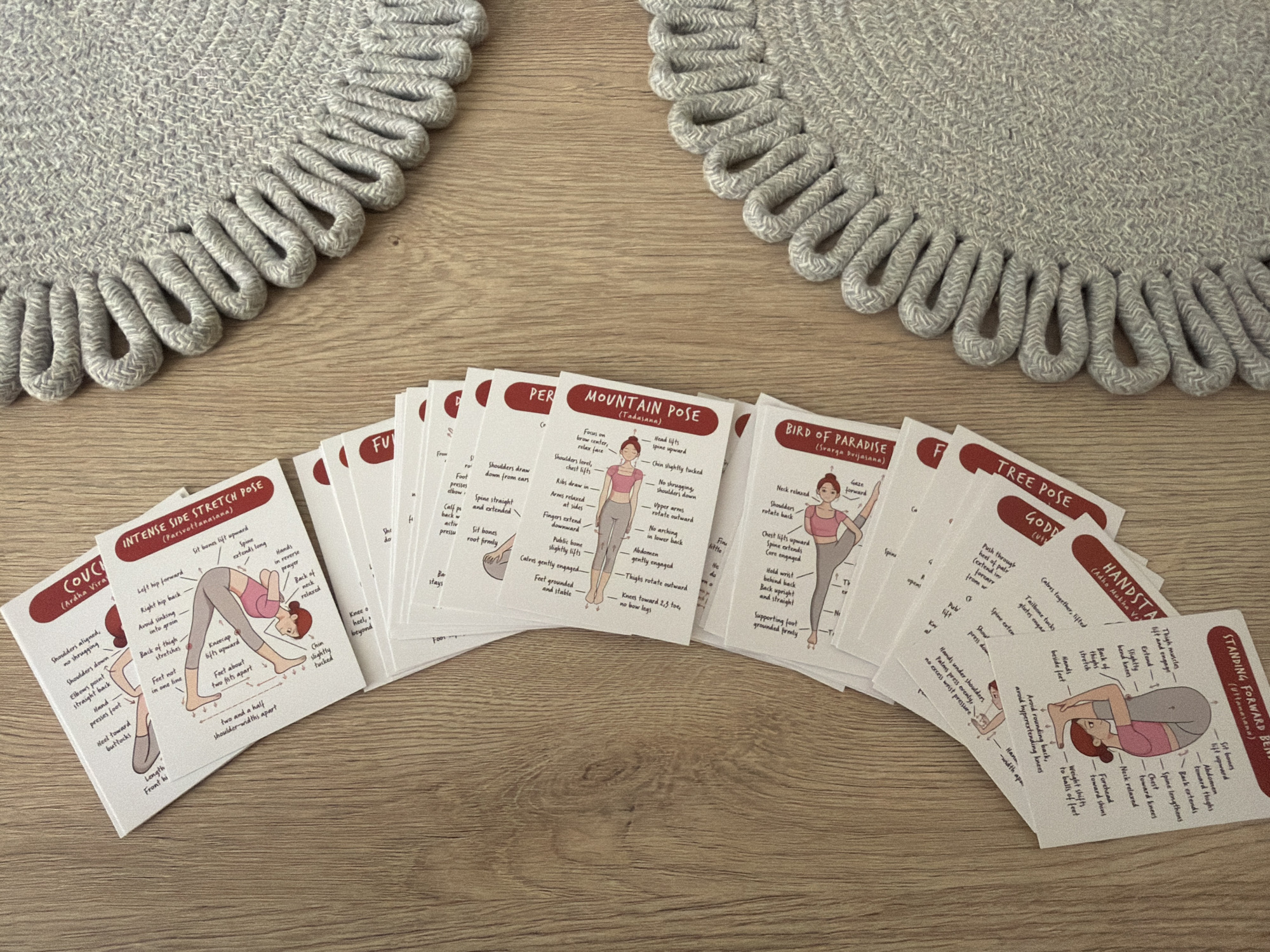 A top-down view shows a collection of illustrated yoga pose cards fanned out on a light brown wooden surface. Two circular, textured grey placemats are partially visible on the right side of the frame, one above the other. The yoga cards, with red titles and black text, depict various poses with instructional diagrams. Examples of visible pose titles include "COUCH (Archa Vira)", "INTENSE SIDE STRETCH POSE (Trikonasana)", "MOUNTAIN POSE (Tadasana)", "BIRD OF PARADISE (Svarga Dvijasana)", "TREE POSE (Vrksasana)", and "HANDSTAND (Adho Mukha Vrksasana)". One card at the bottom is titled "STANDING FORWARD BEND (Uttanasana)". The cards are overlapping and arranged in a semi-circular fanned pattern, suggesting they are laid out for reference or use. The lighting is even, and the focus is sharp on the cards and the wooden surface. There are no people or animals present in the image. The scene is static and domestic.