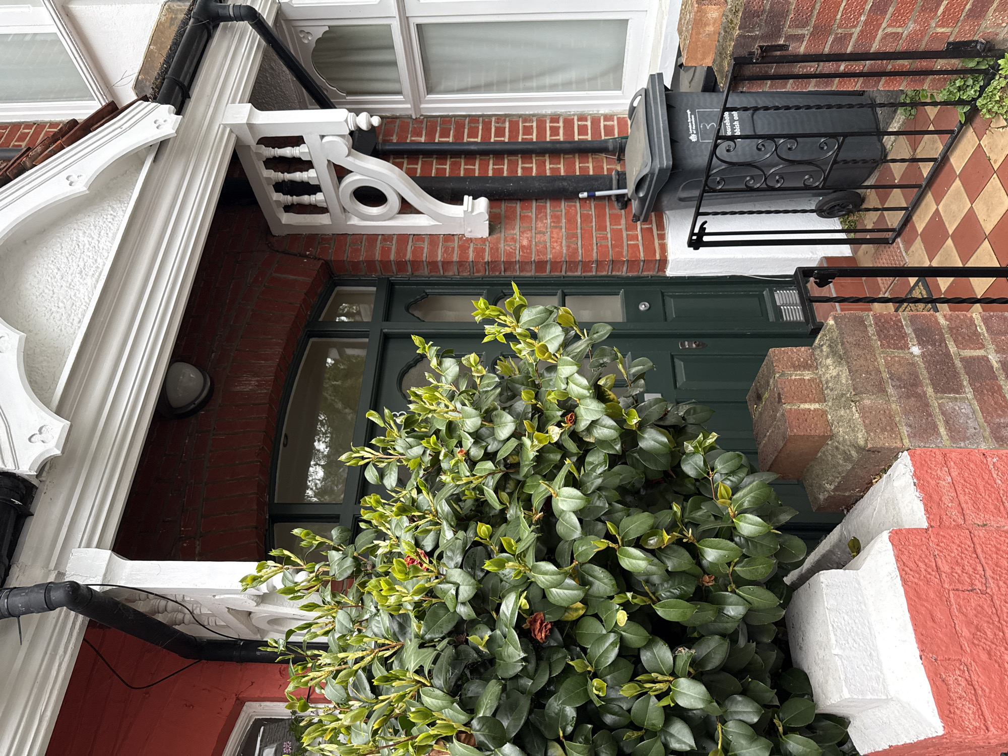 A low-angle, rotated photograph captures a residential facade in London, United Kingdom. The front door is painted dark green, with a window panel above. A large, dense green bush dominates the foreground, partially obscuring the door. Above the door, a black metal bin is mounted on the brick wall, with "London Borough of Hounslow" printed on its side. The building has a mix of red brick, white-painted architectural details, and decorative brackets. A black drainpipe runs along the building. A portion of a window is visible on the upper part of the image. The architecture suggests a typical Victorian or Edwardian terraced house. The image is taken during the daytime.