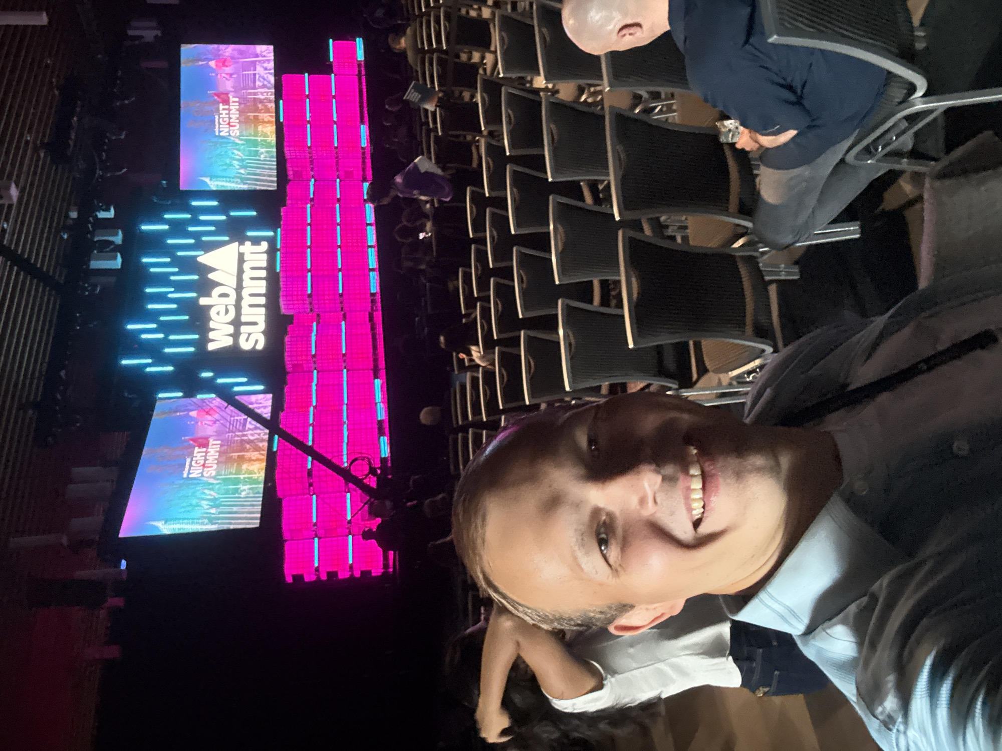 A smiling man in a light blue button-down shirt takes a selfie at the Web Summit conference, as indicated by the illuminated "Web Summit Night Summit" sign in the background. He is surrounded by rows of empty chairs and other attendees. The venue appears to be indoors, possibly a conference hall. There is no evidence to confirm that the image was taken in Vancouver, Canada.