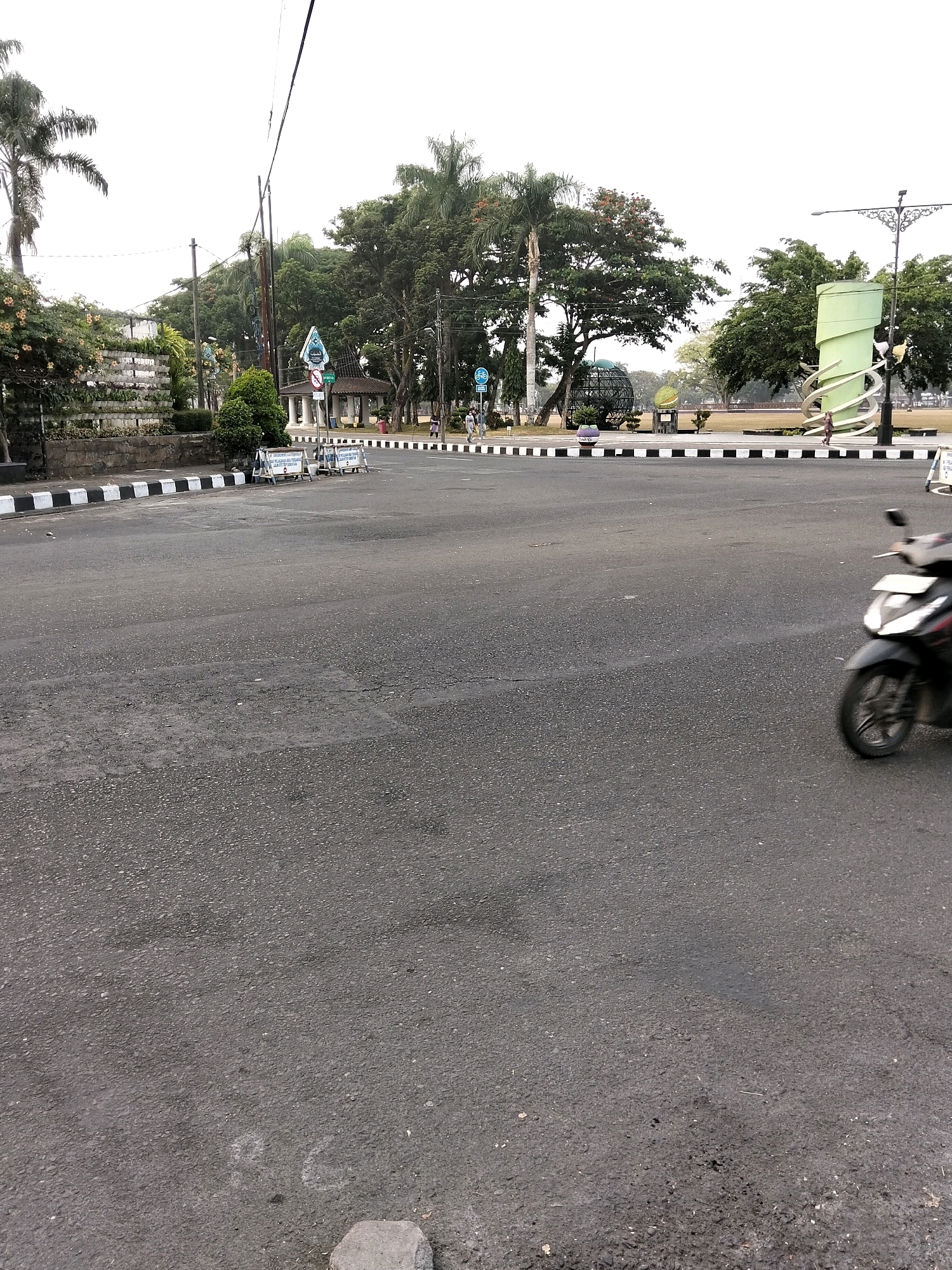 The image captures a view of the alun-alun (town square) in Purworejo, Indonesia, characterized by a large, empty asphalt road in the foreground with a black and white striped curb. In the middle ground, a motorcycle is visible in motion, partially blurred, suggesting movement. The background reveals a park-like setting with trees, a distinctive green spiral sculpture, a geodesic dome structure, and a gazebo. Several people are seen walking in the distance, and temporary barriers or fences are placed along the roadside. The sky is overcast. The overall scene depicts a public space in Purworejo during the day.