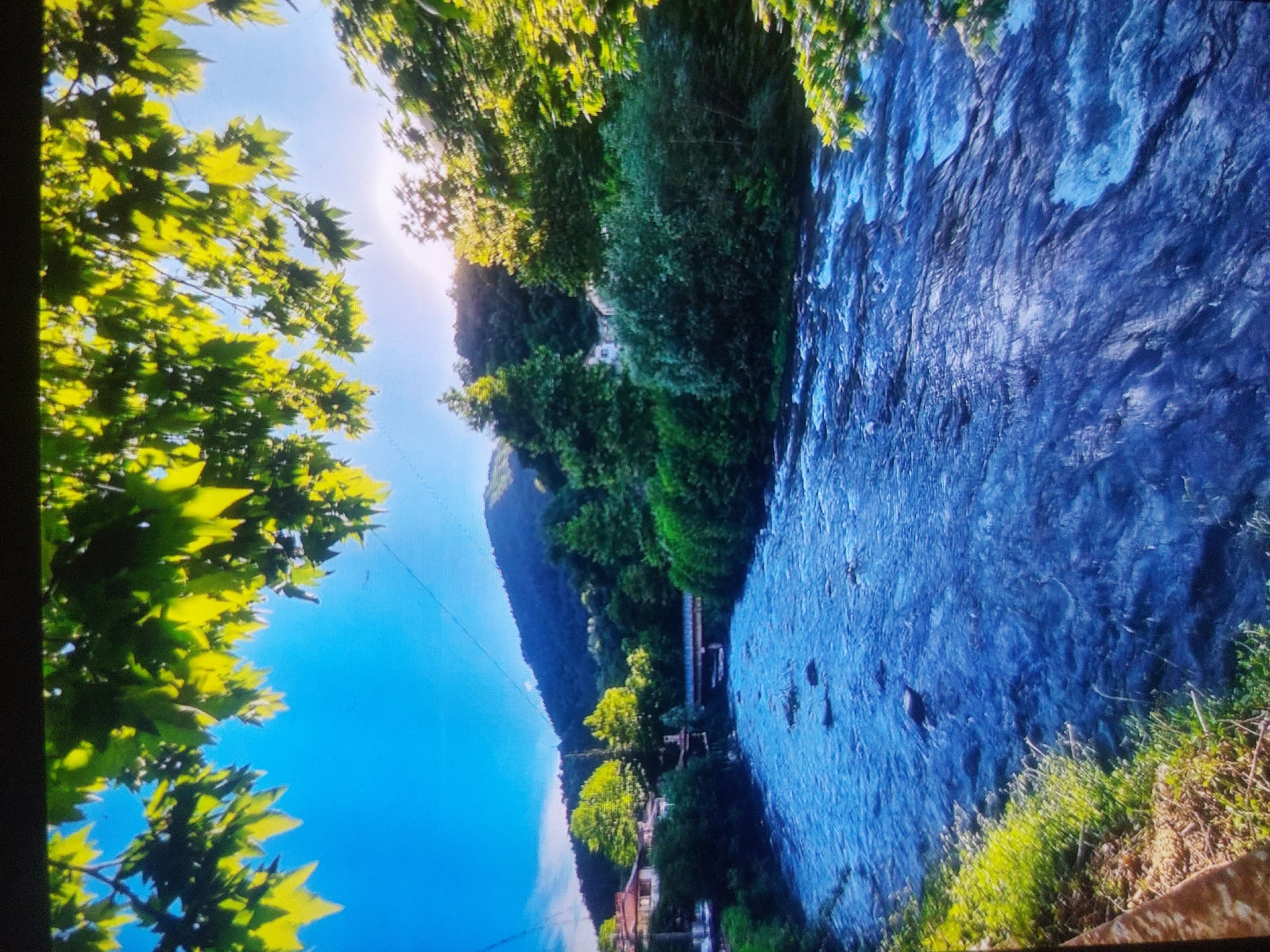 The image shows a river flowing through a landscape with greenery. The river is blue with flowing water, with some rocks visible in the riverbed. Green trees and vegetation line both sides of the river, along with some buildings on the left side of the river. The sky is blue, and the sun shines through the leaves. A power line is visible in the upper left. A mountain is also visible in the background. The image is likely taken in Tsarevo, Bulgaria.