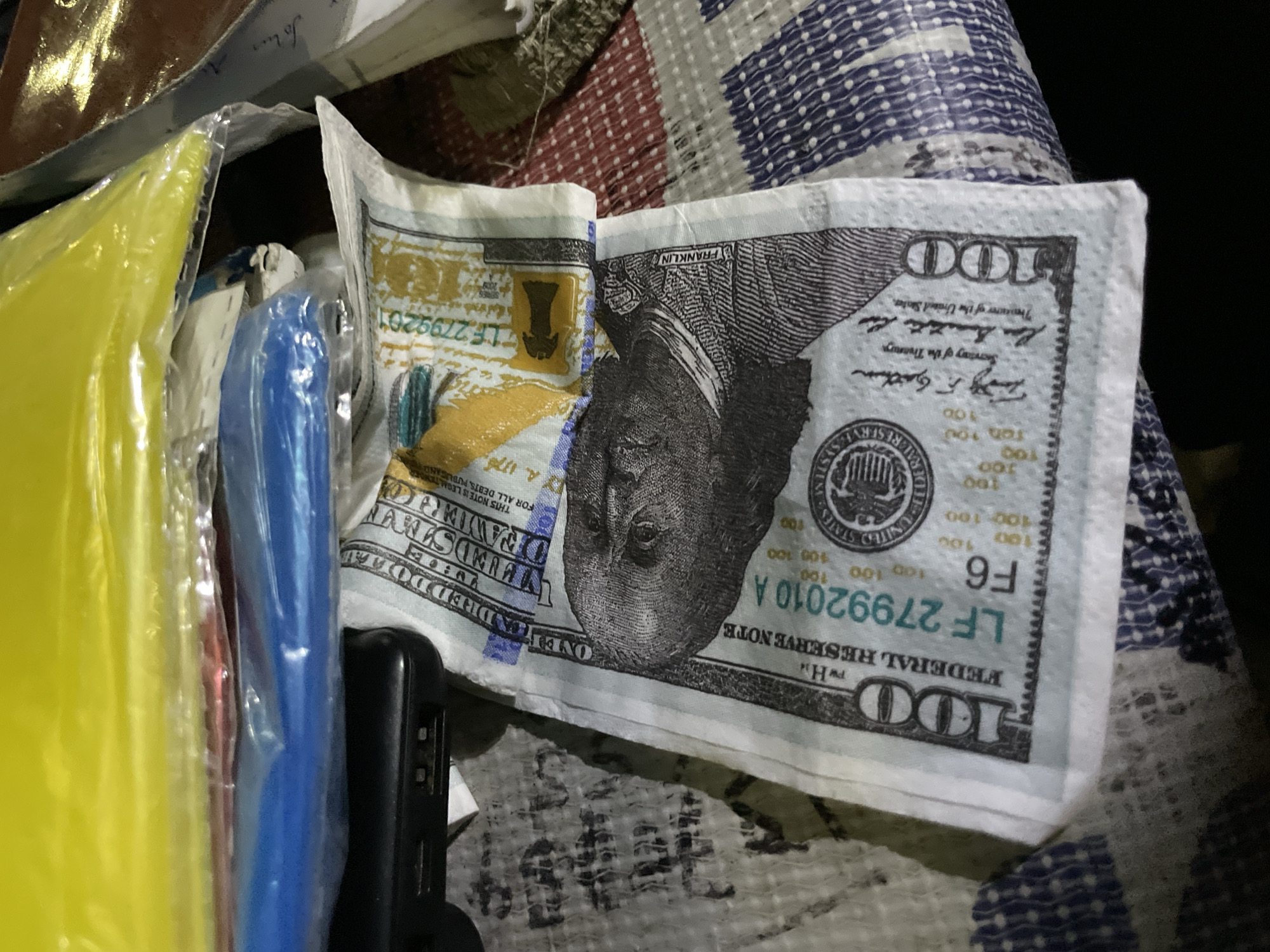 A close-up, overhead shot captures a crumpled one-hundred-dollar bill with the portrait of Benjamin Franklin as the central focus. The bill is partially obscured by other items, including what appears to be a black electronic device with USB ports and several colorful plastic-wrapped objects in shades of yellow, red, and blue, suggesting they might be stationery or other supplies. The textured surface beneath the bill appears to be a patterned fabric, possibly a tablecloth or blanket. The lighting is dim, casting shadows that add to the overall somber tone of the image.
