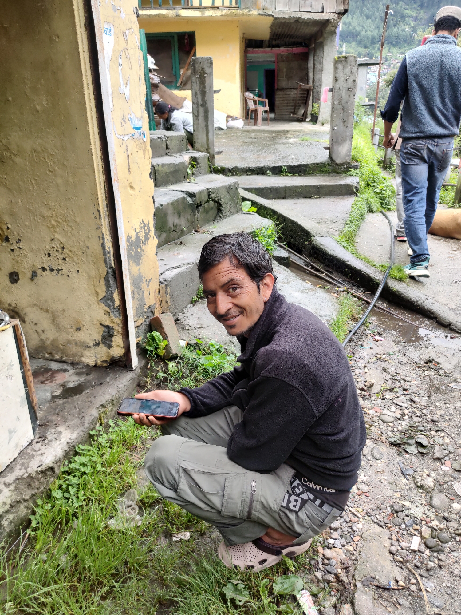 The image depicts a man crouching in front of a weathered yellow building with a textured facade. He is holding a dark-colored smartphone in his right hand. The man is wearing a black fleece jacket and grey cargo pants. His left hand is resting on his knee. He has dark hair and is looking towards the camera with a slight smile. Green grass and some scattered debris, including pebbles and bits of plastic, are visible around him. In the background, concrete stairs lead up to the building, and a partial view of another structure with a green-framed window is visible. Further in the background, a man in a blue sweater and jeans walks away from the camera, and the silhouette of trees and buildings on a hillside suggests a mountainous terrain. The environment appears to be an outdoor, possibly semi-urban or rural setting.