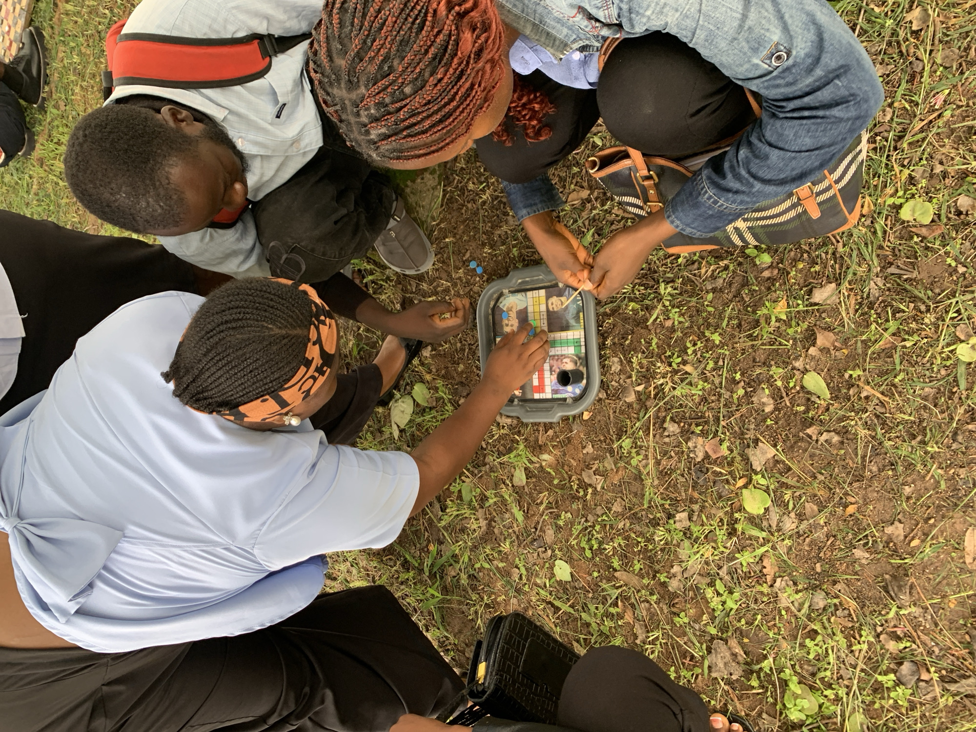 In Rafin Par, Nigeria, a group of individuals are gathered outdoors, seated on the ground, playing a board game. The environment is an unpaved area with sparse grass, dirt, and fallen leaves. The people are dressed in casual clothing. One person, wearing a light blue collared shirt with a bow detail at the back, is leaning over the board game. Another individual, in a light blue shirt and a red backpack, is also engaged in the game. A third person, wearing a denim jacket, has their hands on the game board. A fourth person, dressed in black, is partially visible. The board game itself is contained within a grey plastic tray and features various colored pieces and images of people. The scene appears to be a casual social gathering.