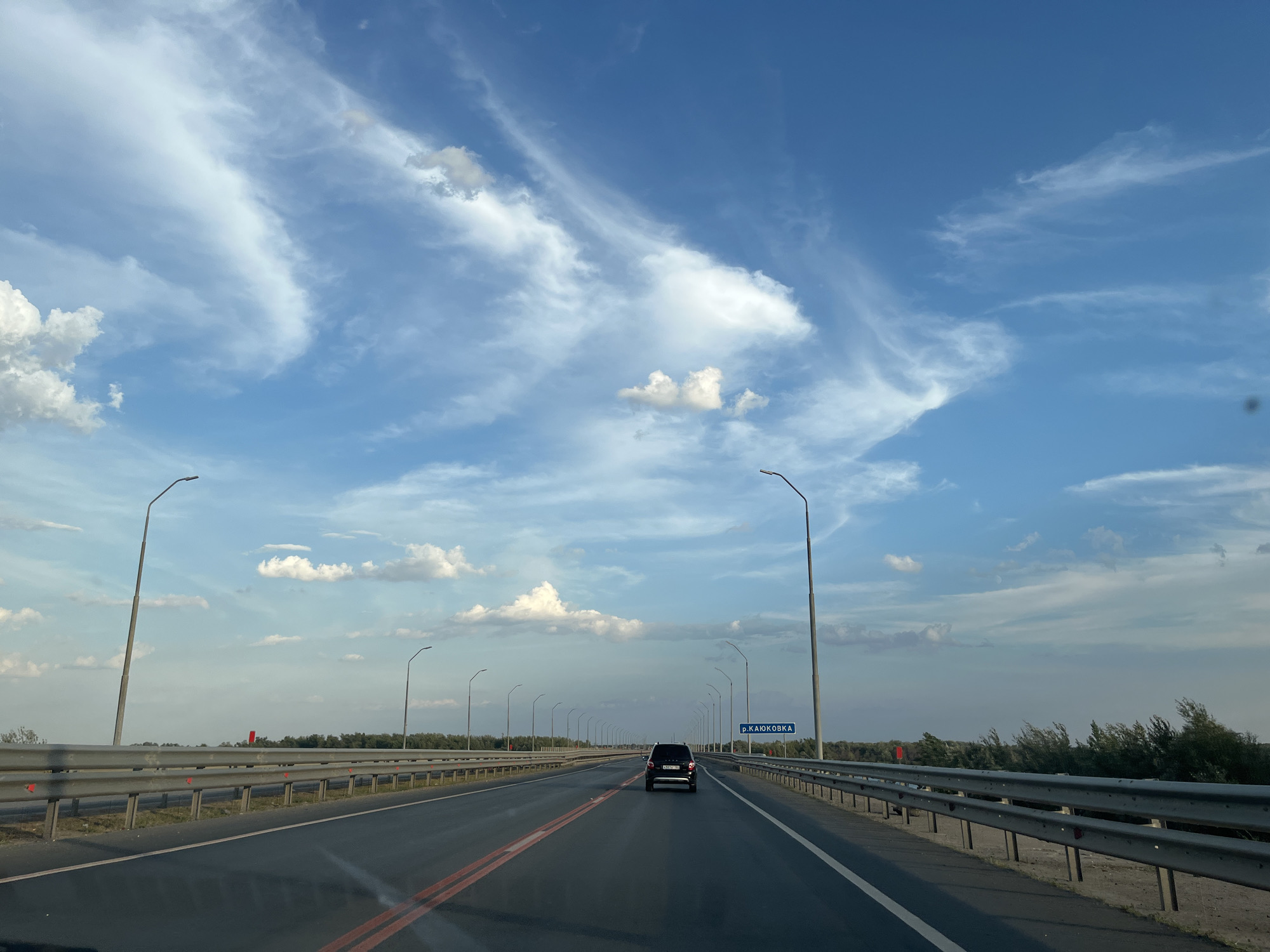 The image shows a black SUV driving on a bridge over a river towards a sign that reads "р. КАЮКОВКА" (River Kayukovks). The bridge is lined with streetlights and a metal guardrail. The sky is blue with scattered clouds. The road has double red lines in the center. The surrounding landscape consists of trees and vegetation on either side of the river. The vehicle appears to be driving in the direction away from the camera. There are no people visible in the image. The scene appears to be a highway or main road in a rural or suburban area.