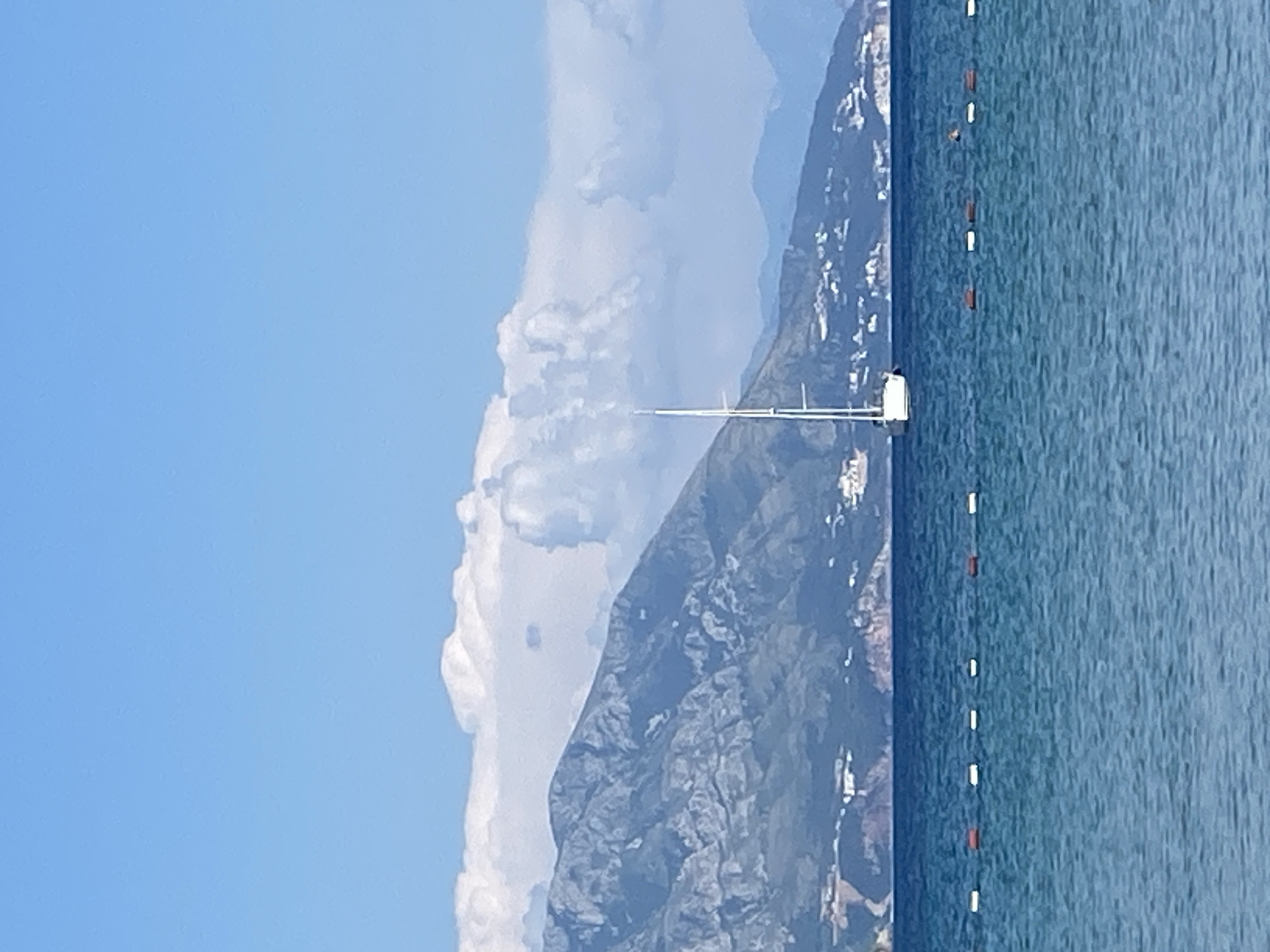 A white sailboat with a tall mast floats on a calm, blue sea. In the background, a rugged mountain range with some scattered white buildings runs horizontally across the frame. Above the mountains, fluffy white clouds drift across a clear blue sky. A rope with alternating white and red buoys marks a boundary in the water in front of the sailboat. The scene appears to be the coast of Budva, Montenegro.
