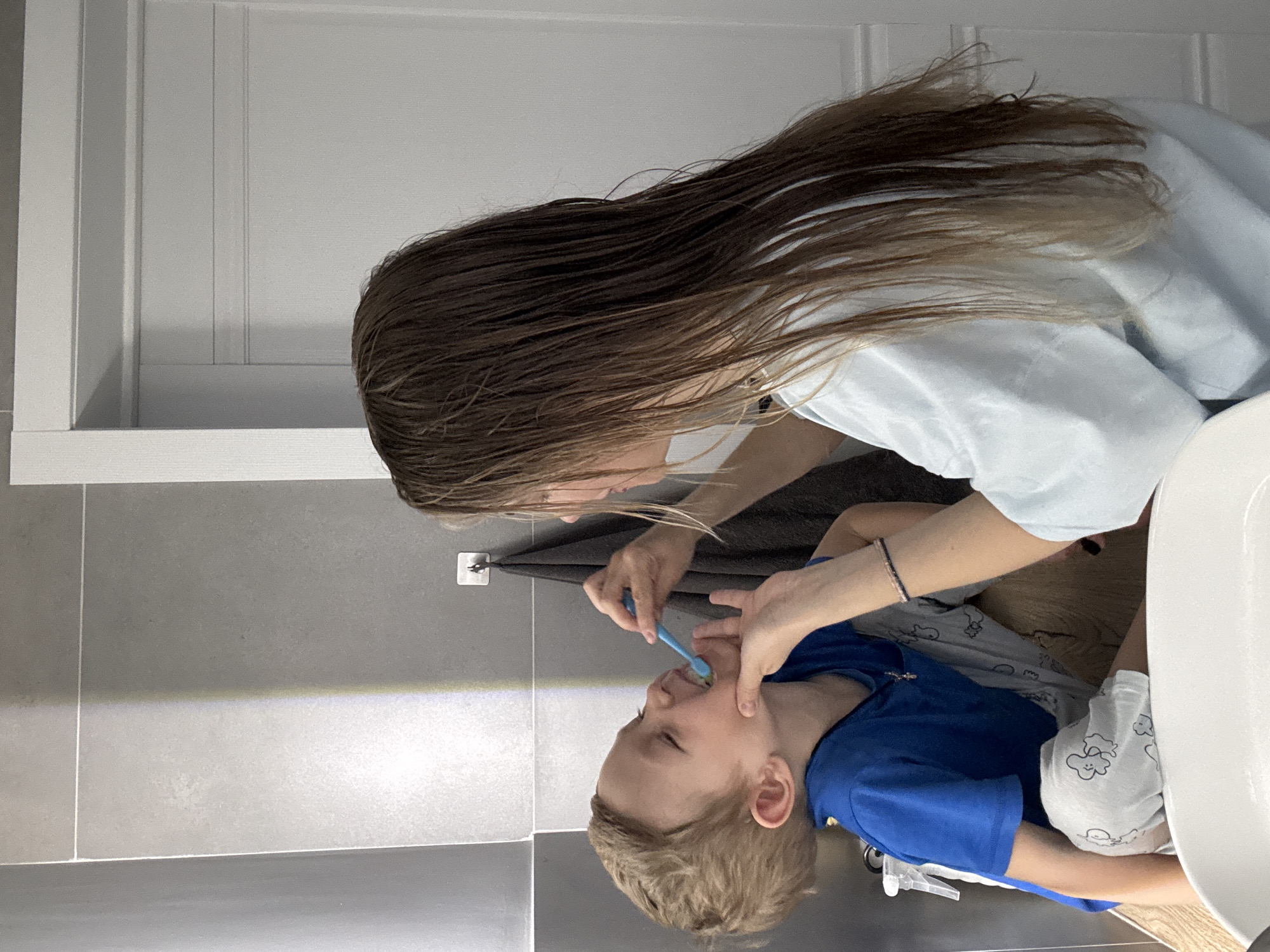 A person with long brown hair is brushing the teeth of a young child. The child is wearing a blue shirt and grey and white patterned shorts. The setting appears to be a bathroom, characterized by grey tiled walls and a white cabinet above the sink. The location is Chaikovsky, Russia.