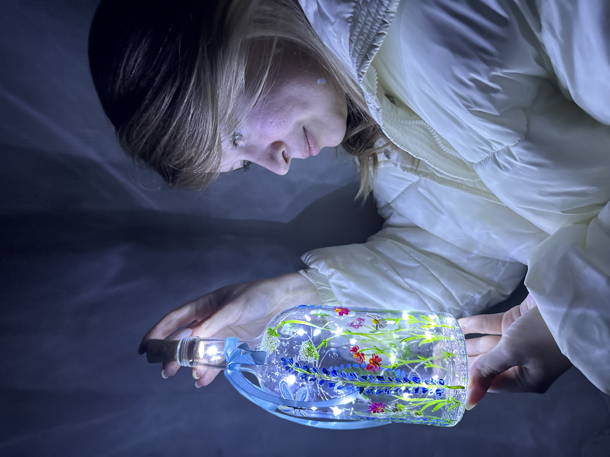 A young woman in a white padded jacket is holding a glass bottle adorned with painted flowers and illuminated by internal fairy lights. The bottle is held horizontally at eye level. The background is dark, suggesting a dimly lit environment. The location, Вассята, Russia, is not visually discernible in the image.