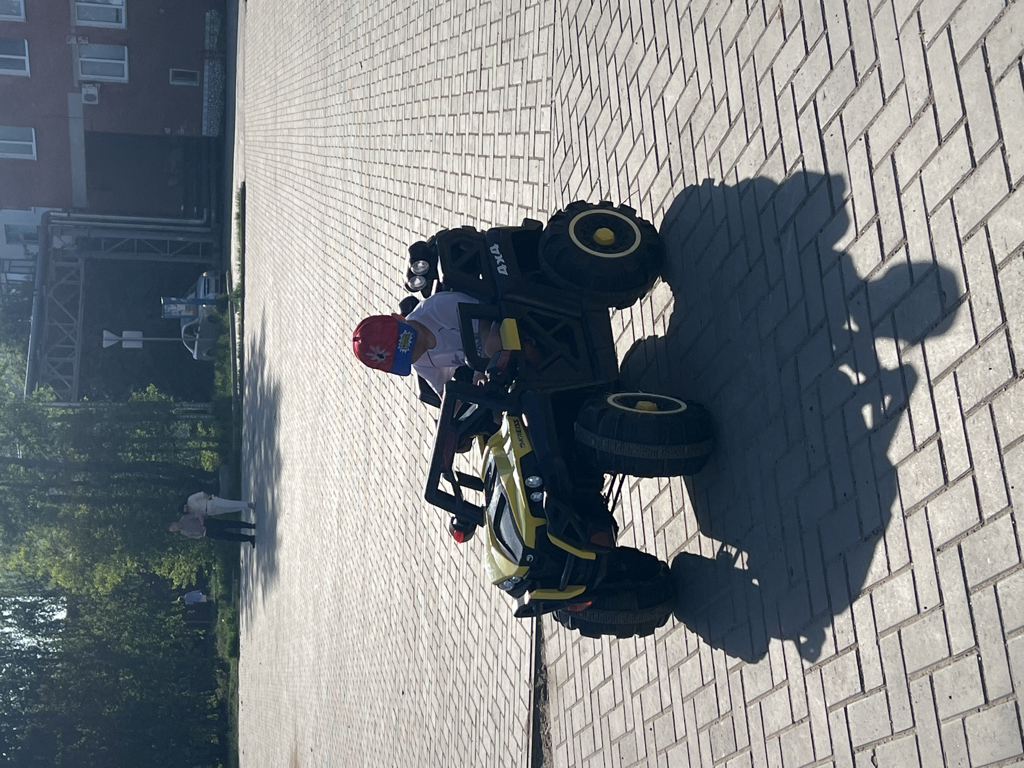 A child is riding a yellow and black four-wheel toy ATV on a gray brick sidewalk in Noviy, Russia. The ATV has large, dark tires with yellow rims and “4X4” written on the side. The child, wearing a red and blue hat, is sitting in the driver's seat, and their shadow is cast on the sidewalk. In the background, there is a building, trees, and two adults.