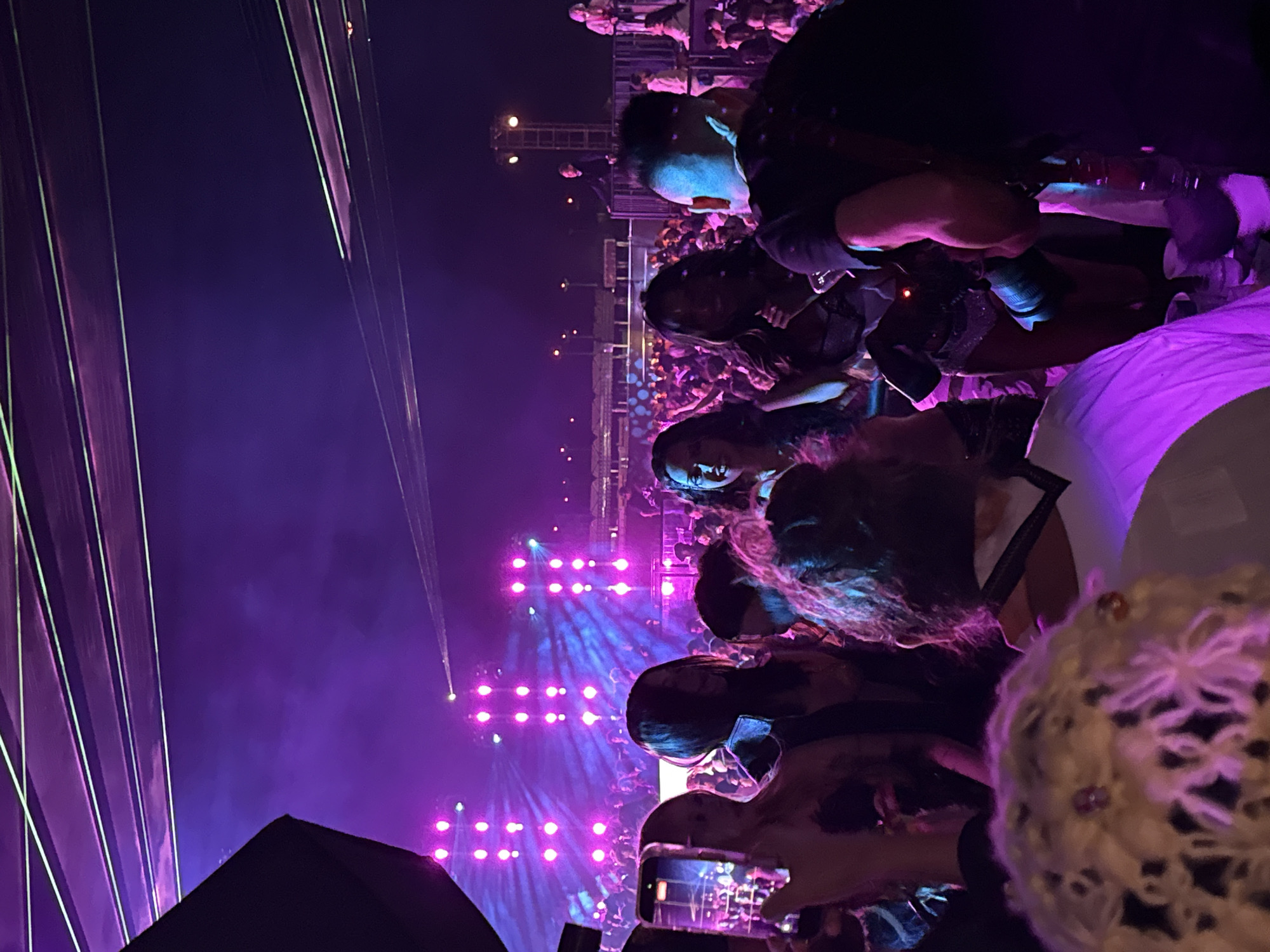 The image depicts a live concert setting, likely at night, in Thermal, United States of America, judging by the context. The foreground shows concert attendees, some with cameras, capturing the event. A person wearing a mask is visible. The background features a large crowd of people watching a performance, highlighted by bright purple and white stage lights and laser beams. A stage or platform structure is visible in the distance, suggesting the location of the performers.