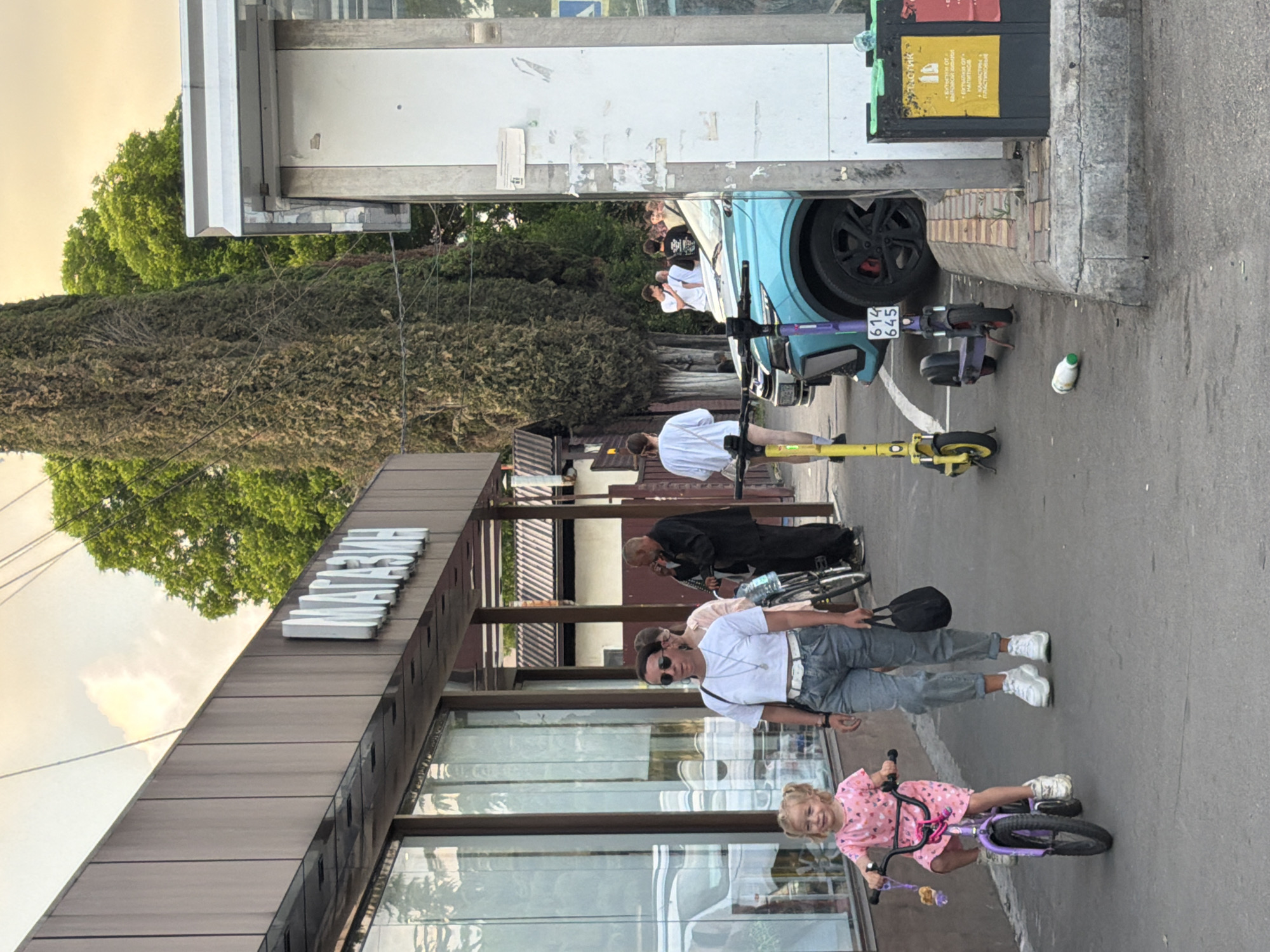 The image shows a group of people and objects on a street, with a building in the background. A young girl on a purple bicycle is in the foreground, smiling. Behind her, a man in a white shirt and jeans stands with his arm around someone. Further back, a group of people are near a light blue car. There are several scooters parked nearby, and a white bottle lies on the ground. The building in the background has a sign that reads "MIRAGE" in large white letters. The scene appears to be outdoors, with trees and a building visible. The lighting suggests it is daytime, possibly late afternoon, with a warm glow in the sky. The overall atmosphere seems casual and relaxed. Some writing is visible on a green bin in the background.