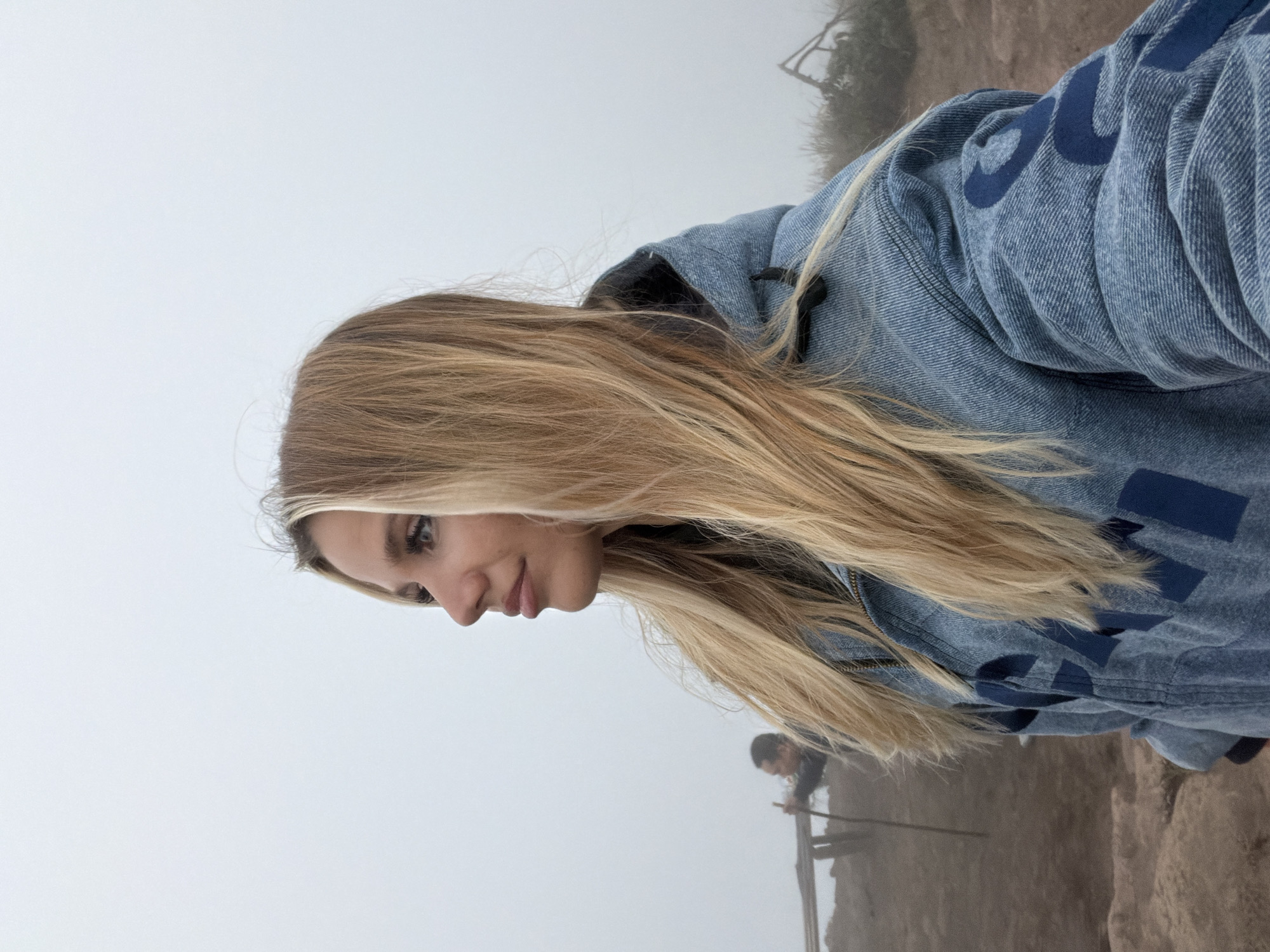 A young blonde woman, facing right in profile, stands in the foreground of an image taken in the city of Kintamani, Indonesia, under a thick, hazy sky. She is wearing a blue denim jacket with a dark hood and large navy blue lettering across the front and sleeves. Her long, blonde hair falls over her shoulders and down her chest. Her eyes are looking upwards and to the right, her lips slightly parted. Her skin is fair and her makeup is subtle, with mascara visible on her lashes.
In the background, a blurred figure of a person holding a stick stands near a wooden structure. The ground appears to be rocky or sandy, with some sparse vegetation visible. The overall atmosphere is misty and damp, indicating a cloudy or foggy day, common in the mountainous region of Kintamani. The scene suggests a moment of observation or contemplation outdoors, likely at an elevated viewpoint overlooking the landscape.