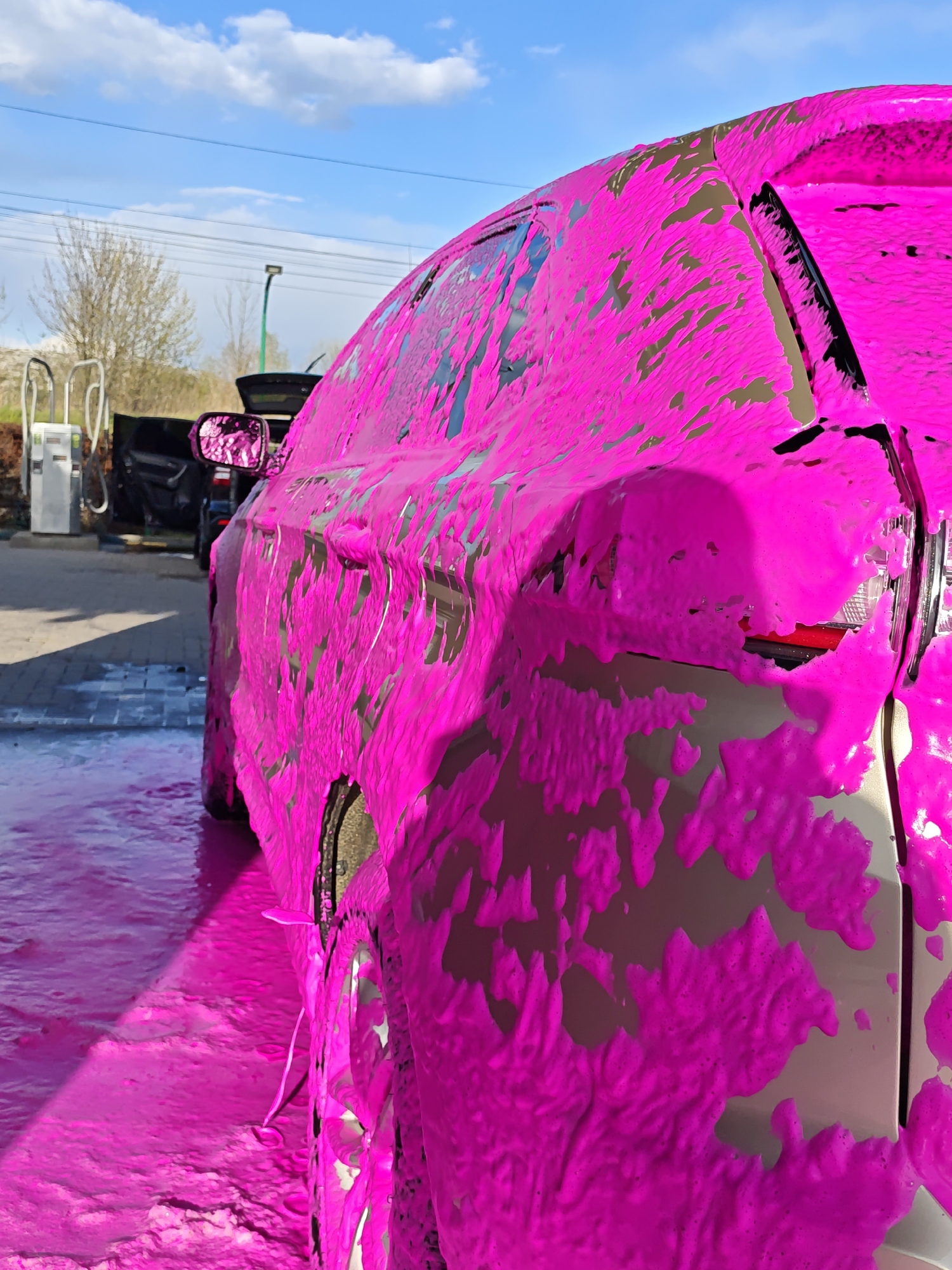 This daytime photograph, taken in Vinnytsia, Ukraine, captures a light-colored car, possibly silver or light gray, at an outdoor self-service car wash. The vehicle is seen from a low angle on its rear passenger side, completely covered in thick, vibrant bright pink cleaning foam. The foam drips down the car's body panels, windows, and partially obscures the rear light and wheels, accumulating on the paved ground below.
In the background on the left, a white self-service car wash unit with hoses or dispensers is visible under a clear blue sky dotted with fluffy white clouds. Behind this equipment, another dark-colored vehicle is partially seen with its trunk open. Bare trees in the distant background suggest an outdoor setting, possibly during early spring. The ground immediately around the car is wet and covered in pink foam residue. The scene vividly depicts the process of a car being washed with a brightly colored cleaning agent. No discernible text is visible on the car or the car wash equipment.