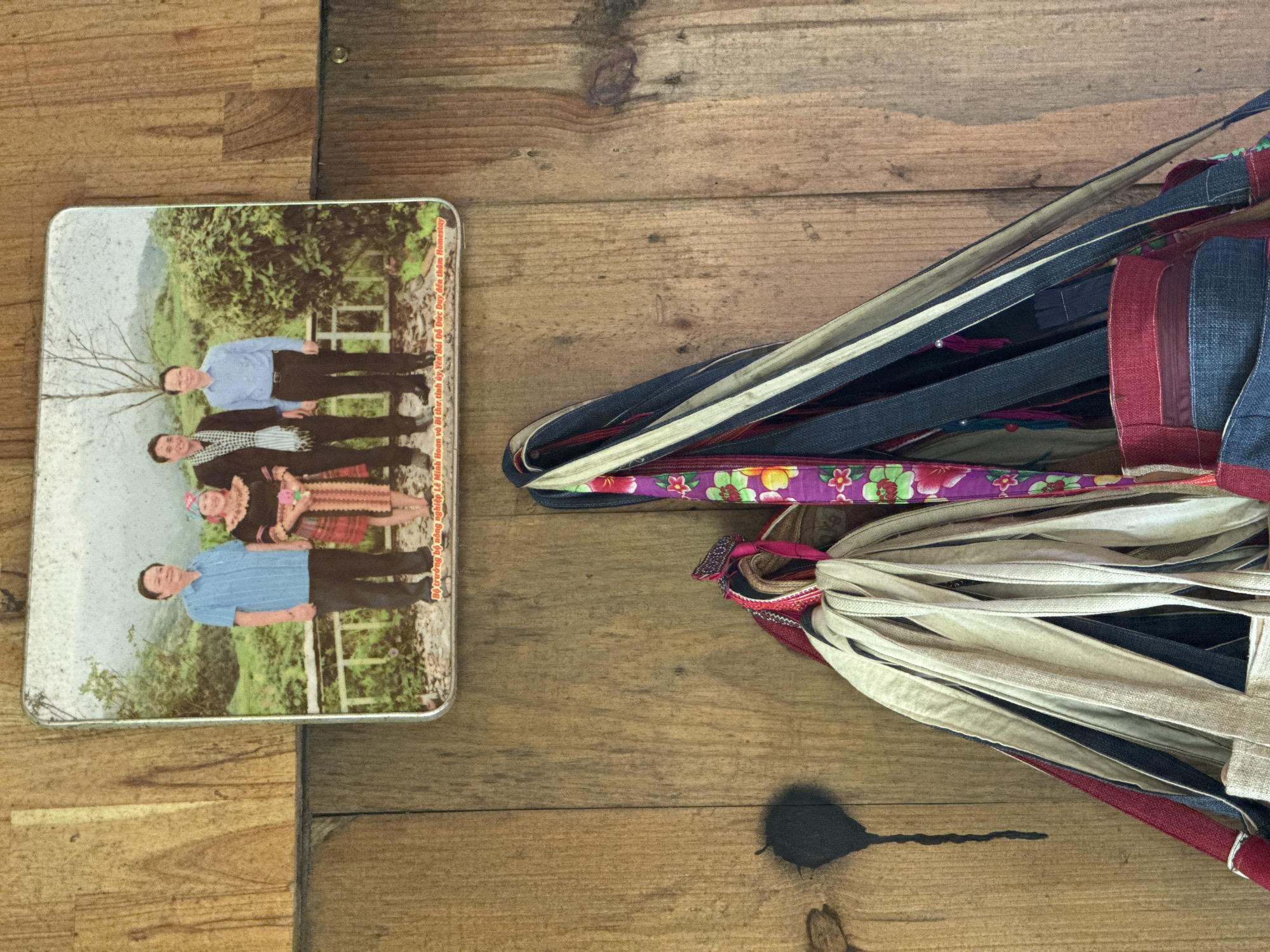 Here is a description of the image:
The image shows a wooden surface with two objects on it: a rectangular metal tin on the left side and a bundle of fabric straps on the right.
The tin features a printed image of a group of people. The people appear to be standing in a landscape with hills and trees. There is text along the right side of the tin which reads, "Bộ trưởng bộ nông nghiệp Lê Minh Hoan và Bí thư tỉnh by Yên Bái Đỗ Đức Duy dán thâm Homestay".
To the right of the tin, a tightly bundled mass of fabric straps is visible. The straps are made from various materials and feature different colors and textures, including red, blue, and tan fabrics. One section of the bundle has a floral pattern. A shadow is cast below the bundle.
The location in the city of Ngã Ba Kim, Vietnam is not determined by the information presented in this image.