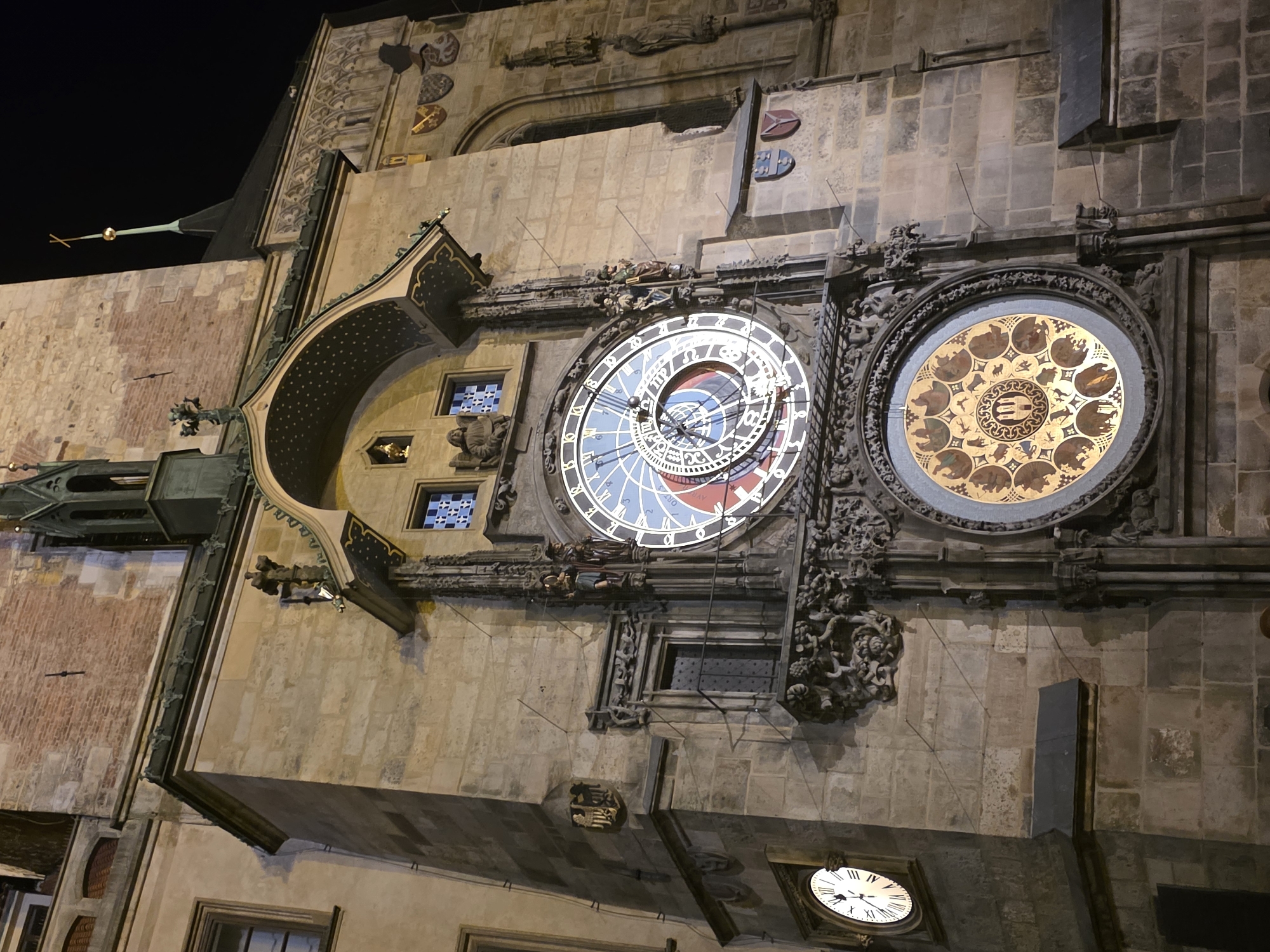 The image captures the Prague Astronomical Clock, a medieval astronomical clock located in the Old Town of Prague. The clock face has a blue and white background with roman numerals and a set of hands indicating the time. Next to the clock is a zodiacal ring. The building's facade is constructed from light brown stone. There are decorative stone carvings and sculptures around the clocks. A smaller clock is visible at the bottom of the image. It is nighttime.