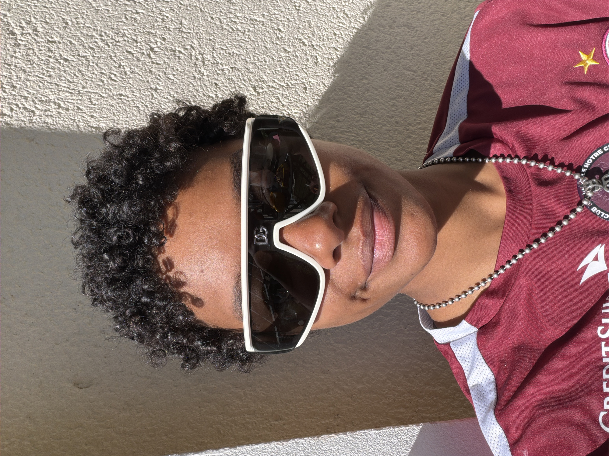 A person wearing a maroon jersey with a white stripe along the sleeve and a silver ball-chain necklace is looking toward the camera and is positioned against a light gray wall with a textured surface. The person is wearing large white-framed sunglasses. The person is wearing a soccer jersey of Angers SCO. The location is Angers, France.