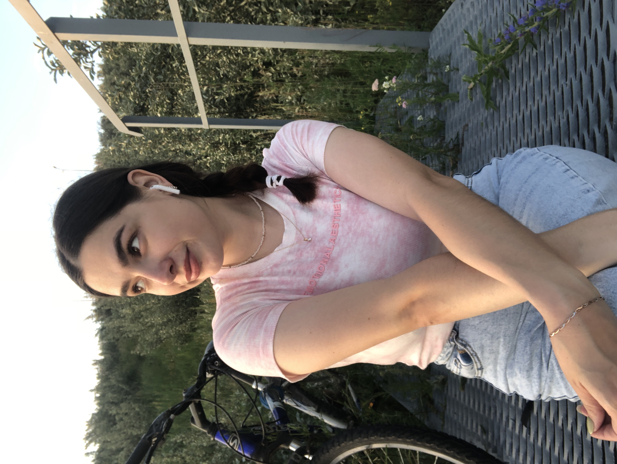 A young woman with dark hair in a braid, wearing a pink tie-dye t-shirt, denim shorts, and a necklace, sits outdoors with her arms crossed. She is wearing white earbuds. To her left, a bicycle is partially visible. She is seated on a metal grate structure, with a background of greenery and a grey metal railing. The image appears to be taken in the city of Chaikovsky, Russia, based on the presence of a specific type of metal grate commonly found in its parks and public spaces, and the overall natural landscape. The lighting suggests it is late afternoon or early evening.