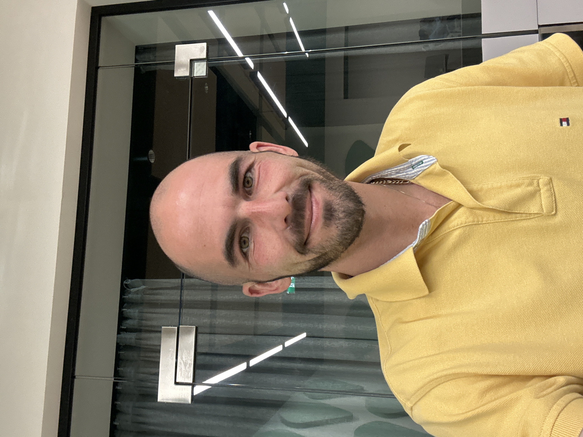 A rotated portrait-style photo shows a man indoors, smiling faintly at the camera. He is bald, with a neatly trimmed dark beard and mustache, and has light brown or hazel eyes. He wears a bright yellow polo shirt with a small, embroidered rectangular logo on the left chest, featuring red, white, and blue sections, reminiscent of the Tommy Hilfiger brand. A gold chain is visible around his neck, partially covered by the open collar of his shirt, which has a blue and white striped lining.
Behind the man is a glass partition or window, reflecting bright, linear lights, suggesting an artificial indoor lighting source. Through the glass, darker, possibly architectural elements and some form of horizontal blinds or patterned surface can be seen. Various metallic fixtures are visible on or through the glass, including a square-shaped one in the upper left background and another resembling an 'L' shape in the lower left. The overall setting appears to be a modern indoor environment, possibly an office or commercial space, in Budva, Montenegro. The man's expression is relaxed and pleasant.