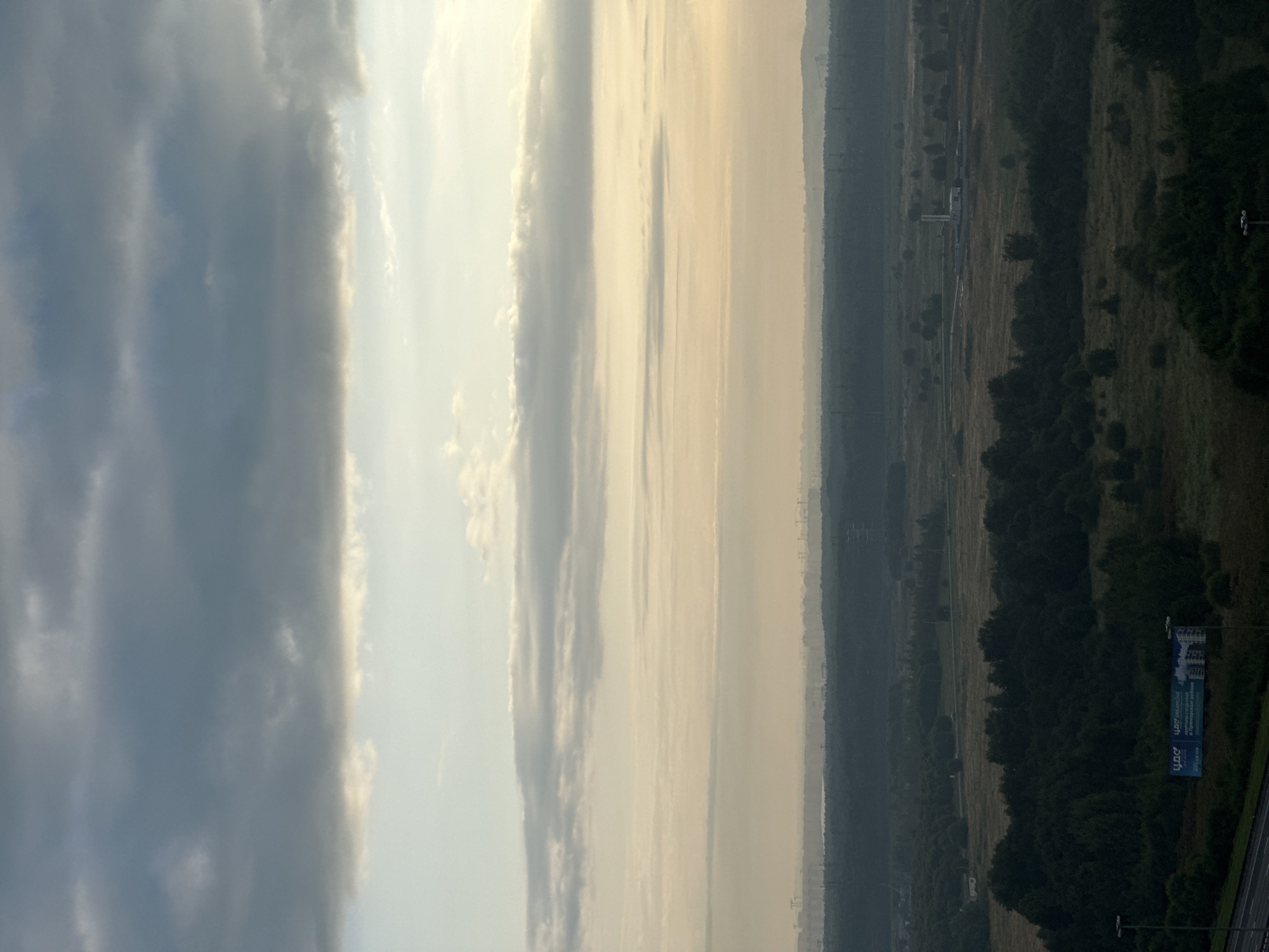 This is an aerial, wide-angle, landscape photograph taken at eye level, facing east. The image depicts a vast expanse of undeveloped land, likely on the outskirts of a city, characterized by rolling hills covered in dense, dark green trees and lighter green meadows. In the distance, a faint urban silhouette can be observed on the horizon, suggesting the presence of a city or town. Several industrial structures, including what appear to be power lines, cranes, and chimneys, are visible amidst the trees and the distant urban landscape. The sky is overcast, with multiple layers of clouds in varying shades of gray and white, and a soft, diffused light suggests it may be early morning or late afternoon. In the foreground, a small section of a dark-colored road is visible on the left, with a street lamp and a billboard advertising an apartment complex in Primorsky District. The overall atmosphere is serene and somewhat muted, with a focus on the natural landscape and the distant signs of human activity. Based on the context provided, this scene is located in Lednevo, Russia.