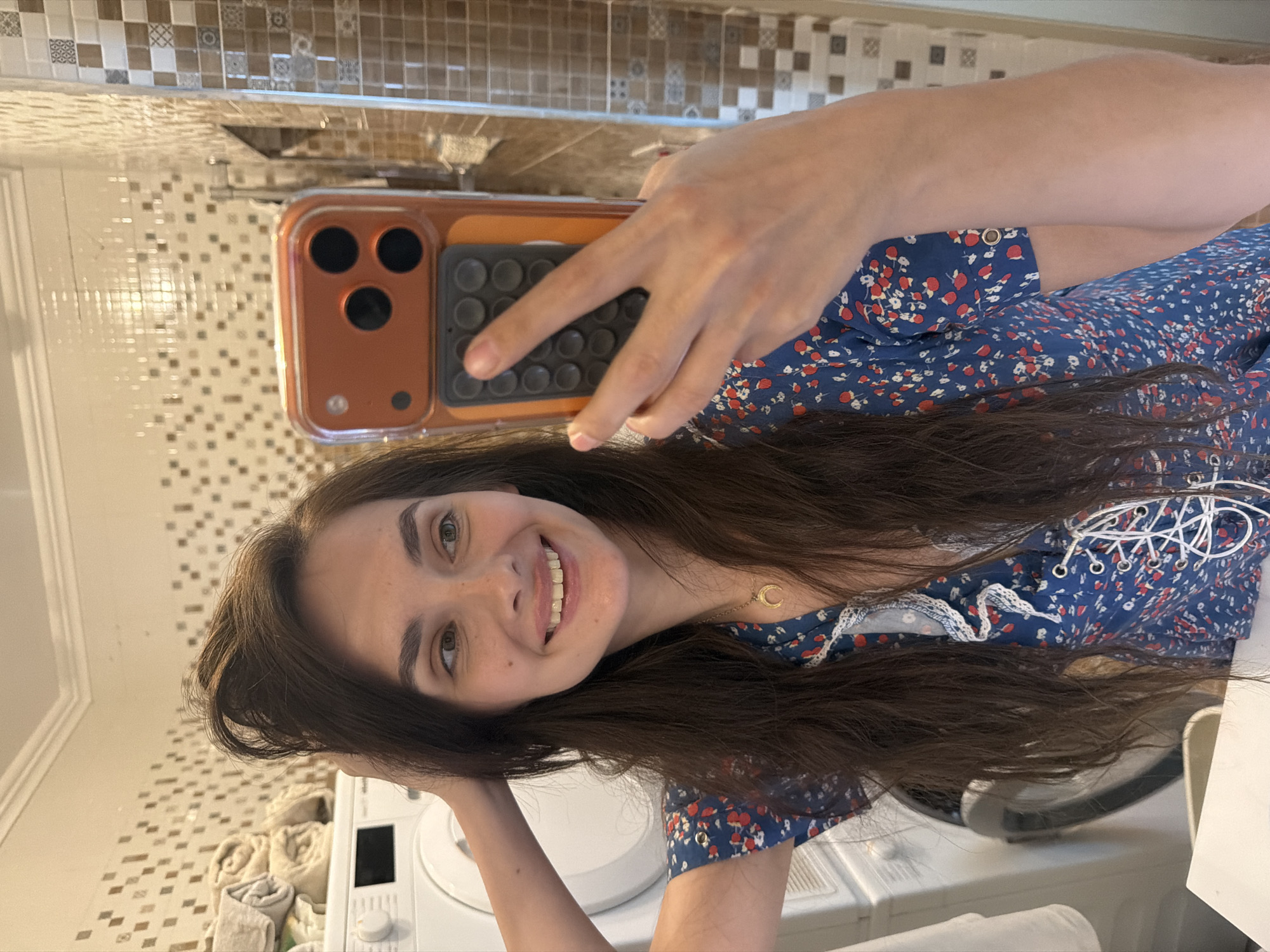 This is a vertical, slightly blurry, full-body selfie taken by a young woman in a bathroom. She is wearing a blue floral dress with white trim and lacing. The woman has long, dark brown hair and is smiling at the camera. She is holding a phone with an orange case, which she is using to take the picture.
The background of the photo shows a tiled wall, a washing machine, and a stack of rolled-up towels. The lighting suggests it is daytime. The overall mood is casual and happy. There is no visible text in the image.