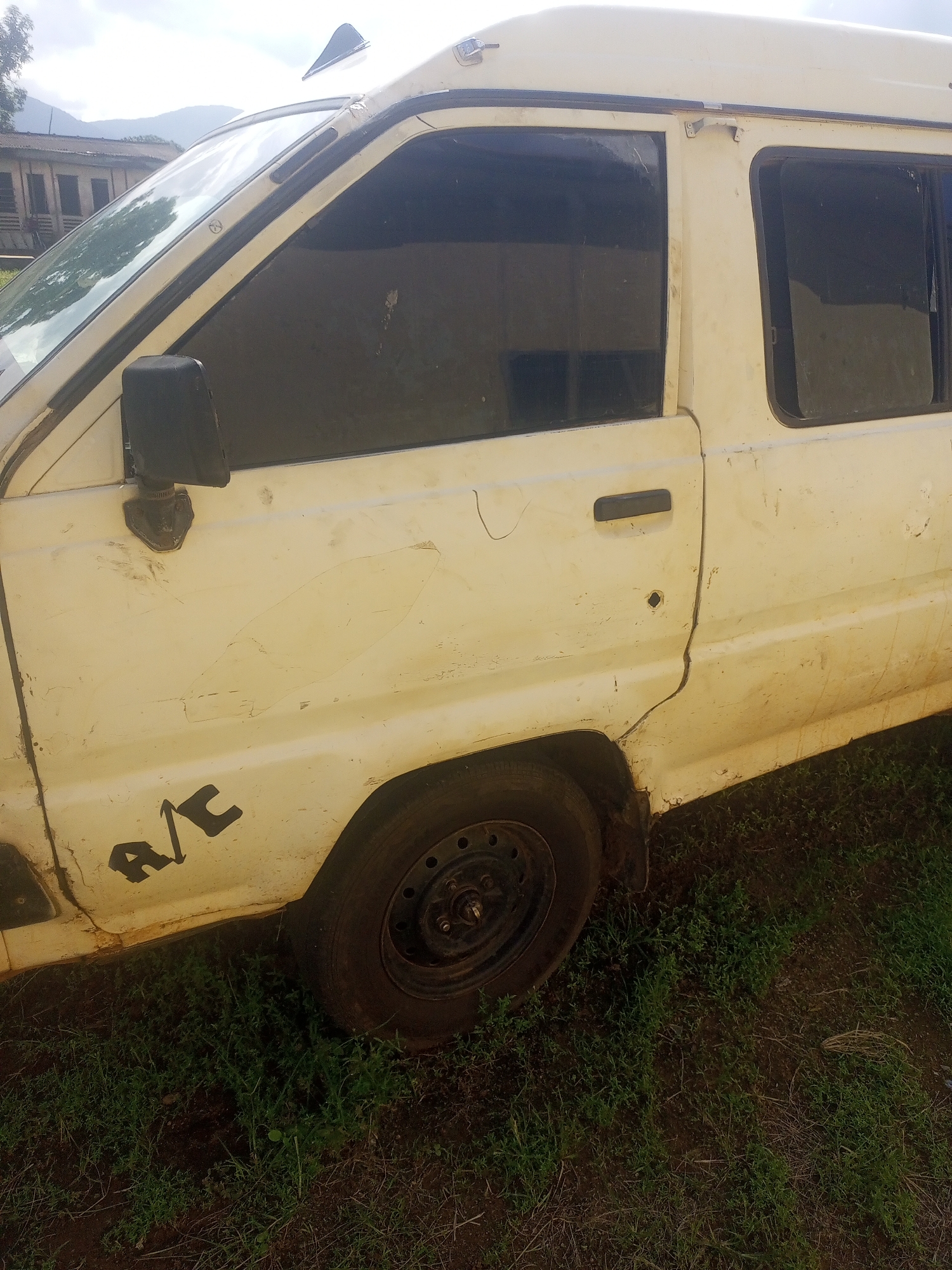 The image shows the side of a light-colored van parked on unpaved ground with sparse green vegetation. The van is dirty and appears to be in disrepair, with visible scratches and dents on its body. The front passenger door is visible, along with the side mirror and the "A/C" decal on the lower part of the door. A spare tire is mounted on the ground near the rear of the visible portion of the van. In the background, a building with a red roof is visible, situated behind some trees and hills. The sky is overcast. This scene is located in Serti, Nigeria.