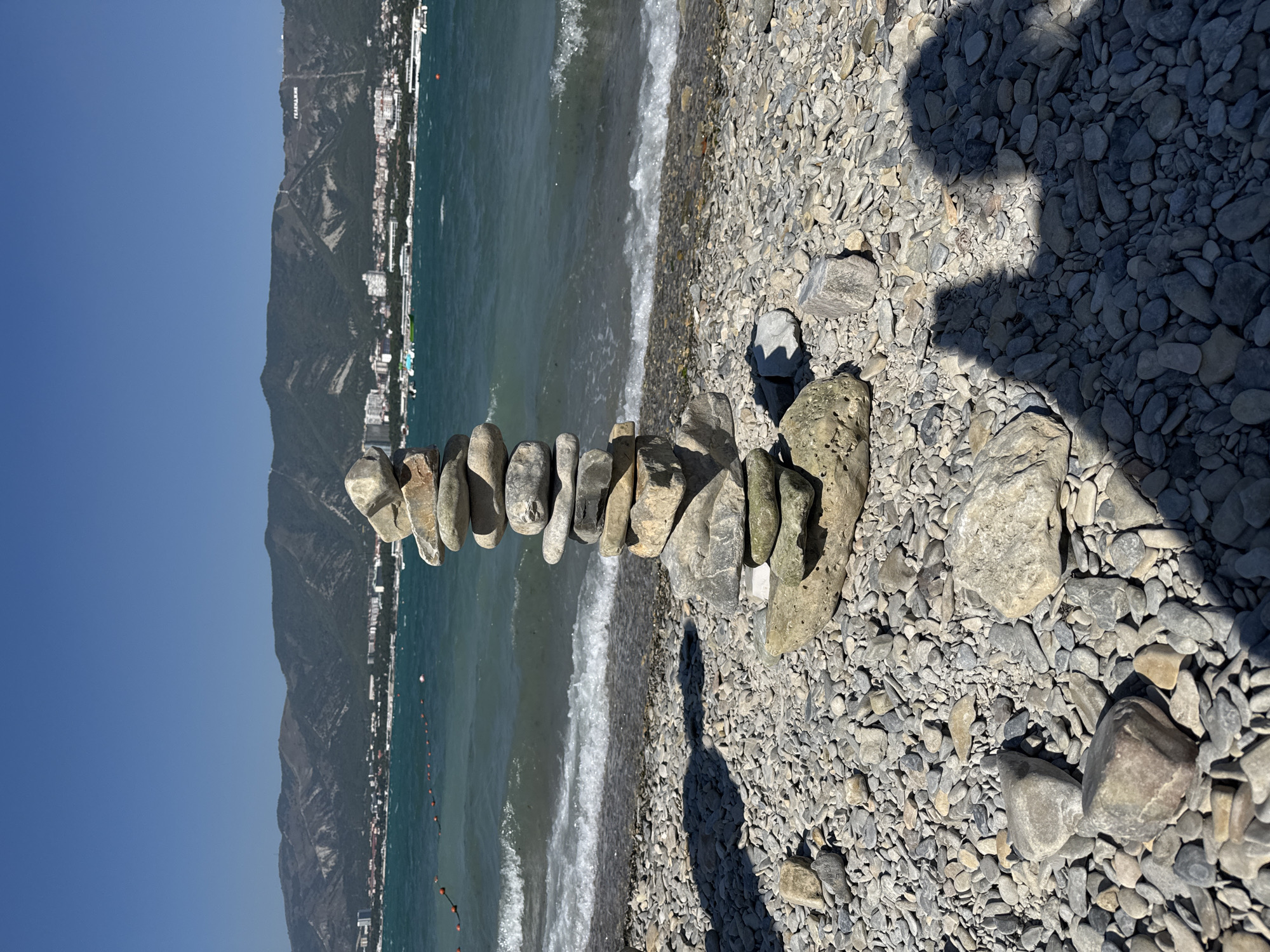 A vertical, medium shot captures a stack of weathered, grey stones meticulously balanced on a pebble beach. The stones are arranged in a tapering tower, with flatter, larger stones forming the base and smaller, more rounded stones at the top. The beach is covered in an expanse of small, grey pebbles, interspersed with larger rocks. In the background, the turquoise waters of the sea gently lap against the shore, creating a foamy white line of waves. Beyond the sea, a city with white buildings and a backdrop of lush, green mountains is visible. A white inscription, reading "ГЕЛЕНДЖИК" (Gelendzhik), is prominently displayed on the mountainside, identifying the location. The sky is a clear, cloudless blue. The foreground of the image is partially obscured by the dark shadows of two individuals, suggesting their presence just outside the frame.