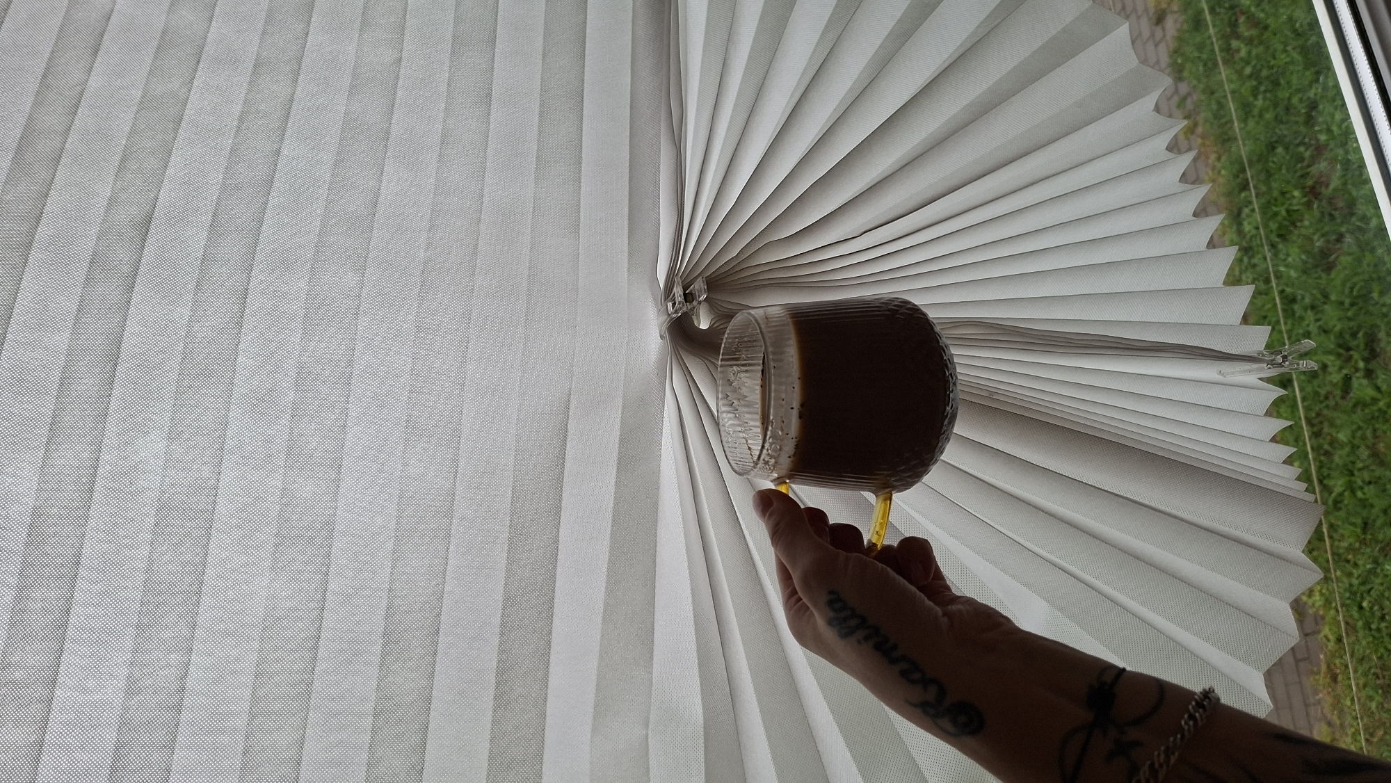 A close-up, eye-level, medium shot shows a person's left arm and hand holding a clear glass mug filled with coffee. The mug has a ribbed texture and a yellow handle. The hand has tattoos on the forearm, including the word "Camilla" written in script. The person is standing near a window with white pleated blinds that are partially open, creating a fan-like effect. Through the gap in the blinds, a glimpse of green grass and a brick walkway is visible. The scene is indoors, with the lighting suggesting daylight. The location is identified as Myadzyel, Belarus.