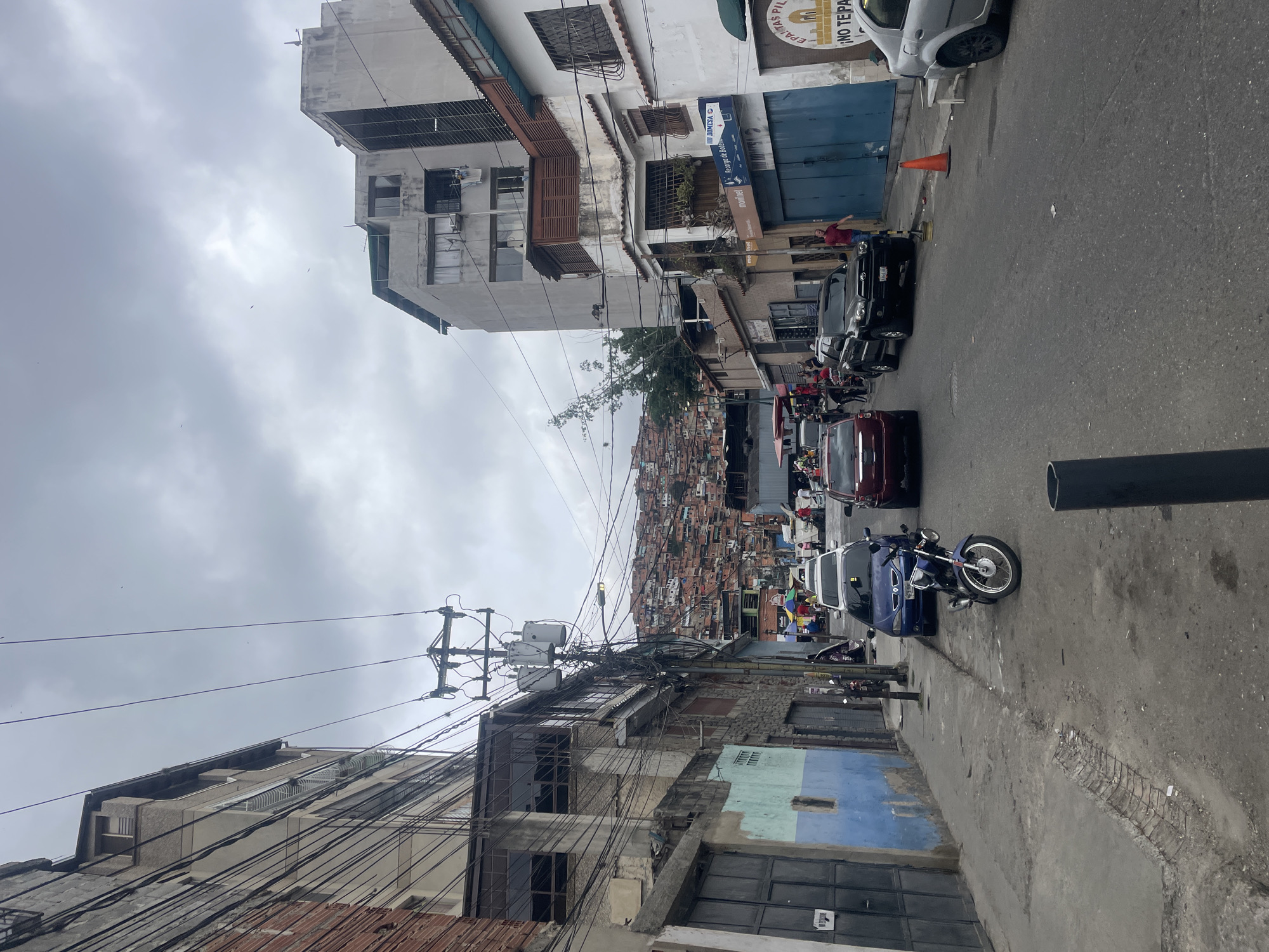 The image depicts a street scene in Caracas, Venezuela. The street is lined with buildings of varying heights and conditions, some appearing dilapidated and others more modern. A dense cluster of small, informal houses covers the hillside in the distance. Power lines are strung haphazardly across the street, creating a cluttered visual effect. Vehicles, including cars and motorcycles, are parked along the road. Some individuals are visible on the street and sidewalk. The sky is overcast, suggesting a cloudy day. The overall impression is of a densely populated urban area with a mix of formal and informal development.
