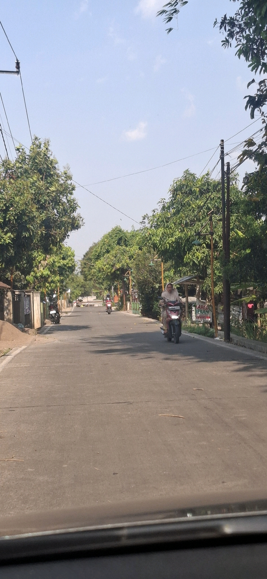 The image depicts a street scene in Cibinong Hilir, Indonesia, on a clear, sunny day. The road is paved and appears to be two lanes wide. Lush green trees line both sides of the street, casting shadows on the asphalt. Overhead, power lines are visible, supported by utility poles.
In the foreground, the bottom edge of what appears to be a car's windshield or dashboard is visible, suggesting the photo was taken from inside a vehicle.
Further down the road, a woman wearing a hijab is riding a red scooter. She is positioned in the right lane, moving away from the camera. Another person on a red scooter is visible further down the road in the left lane. In the distance, more trees and buildings are faintly visible, indicating a residential or mixed-use area.
On the left side of the road, there is a low wall and a pile of sand or dirt. On the right side, there are utility poles, some street lamps, and a sign with red and white lettering that is partially obscured. The overall atmosphere is that of a typical day in a suburban Indonesian town.