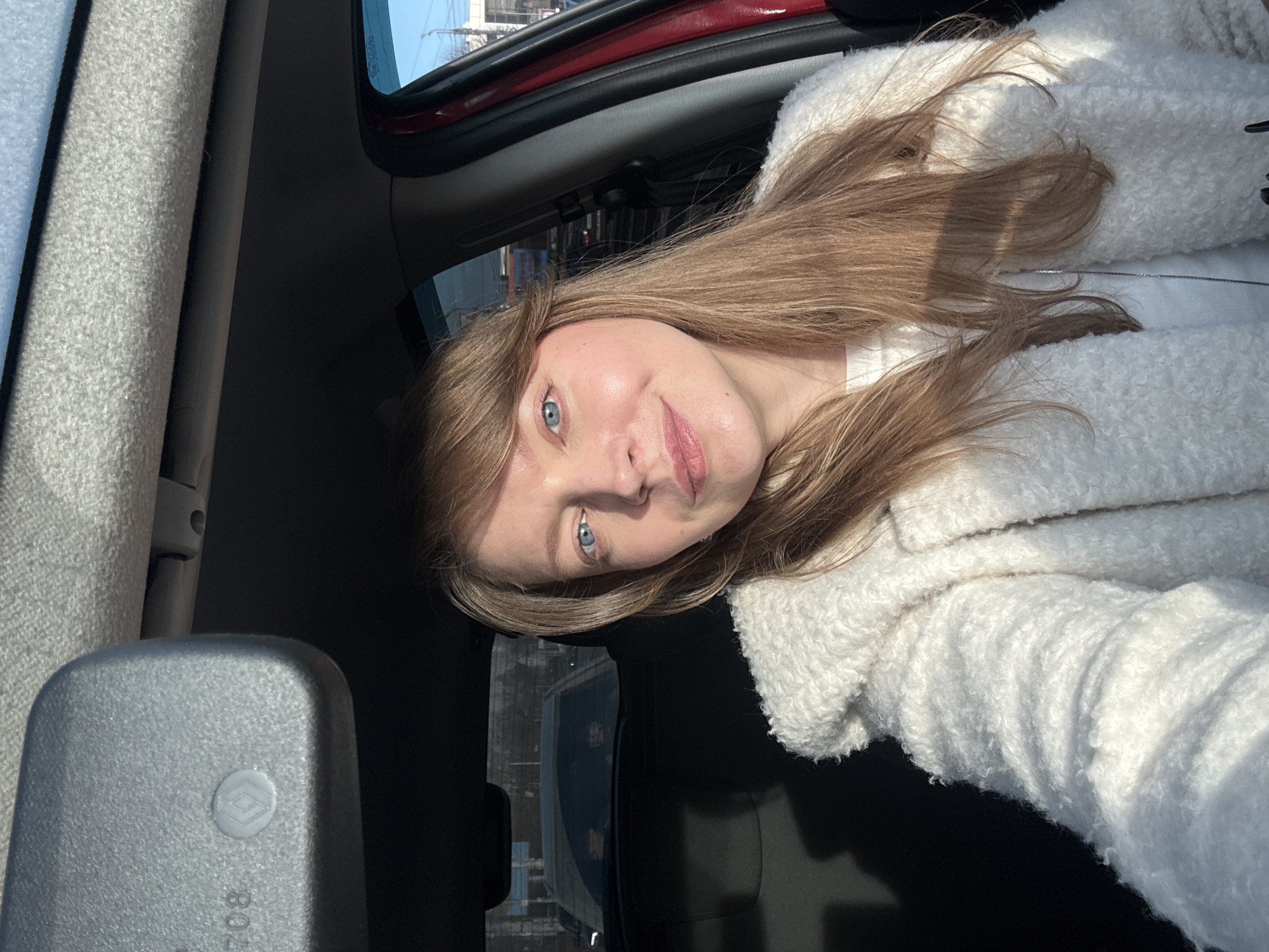 The image displays a blonde woman inside a stationary vehicle, taking a selfie. She is positioned in the front driver's or passenger seat, looking directly at the camera with a subtle smile. She has light brown, shoulder-length hair with bangs, blue eyes, and wears a white, textured coat over a white garment. Direct sunlight illuminates her face and the right side of her body.
The vehicle's interior includes a dark gray rearview mirror in the upper left, featuring an embossed Renault logo and a faint, reversed number "708" on its surface. The headliner is light gray and textured. A red car door panel or pillar is visible along the right edge of the frame. Through the rear windshield, another vehicle is visible, followed by a blurry urban background. The side window on the right reveals a bright blue sky above indistinct urban structures. The scene is consistent with an urban environment within Izhevsk, Russia, but specific street names or landmarks are not discernible from the provided visual information.