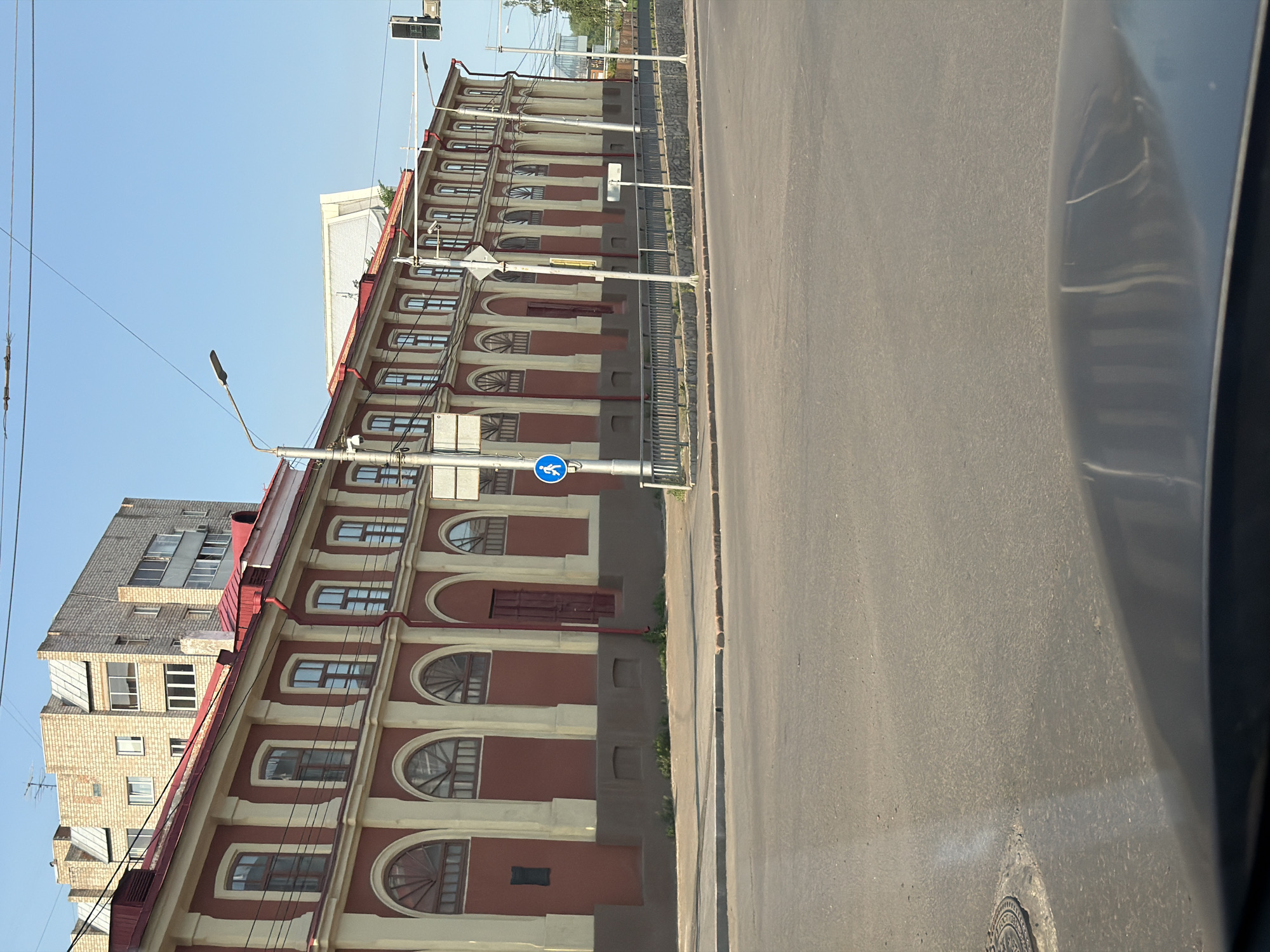 The image shows a street scene in Krasnoyarsk, Russia, featuring a long, red and cream-colored building with arched windows. A modern, light-colored building is visible behind it. The street in the foreground is paved, with a manhole cover visible on the left. A pedestrian crossing sign is posted on a pole to the right of the building. Overhead wires and streetlights are also present. The sky is clear and blue.