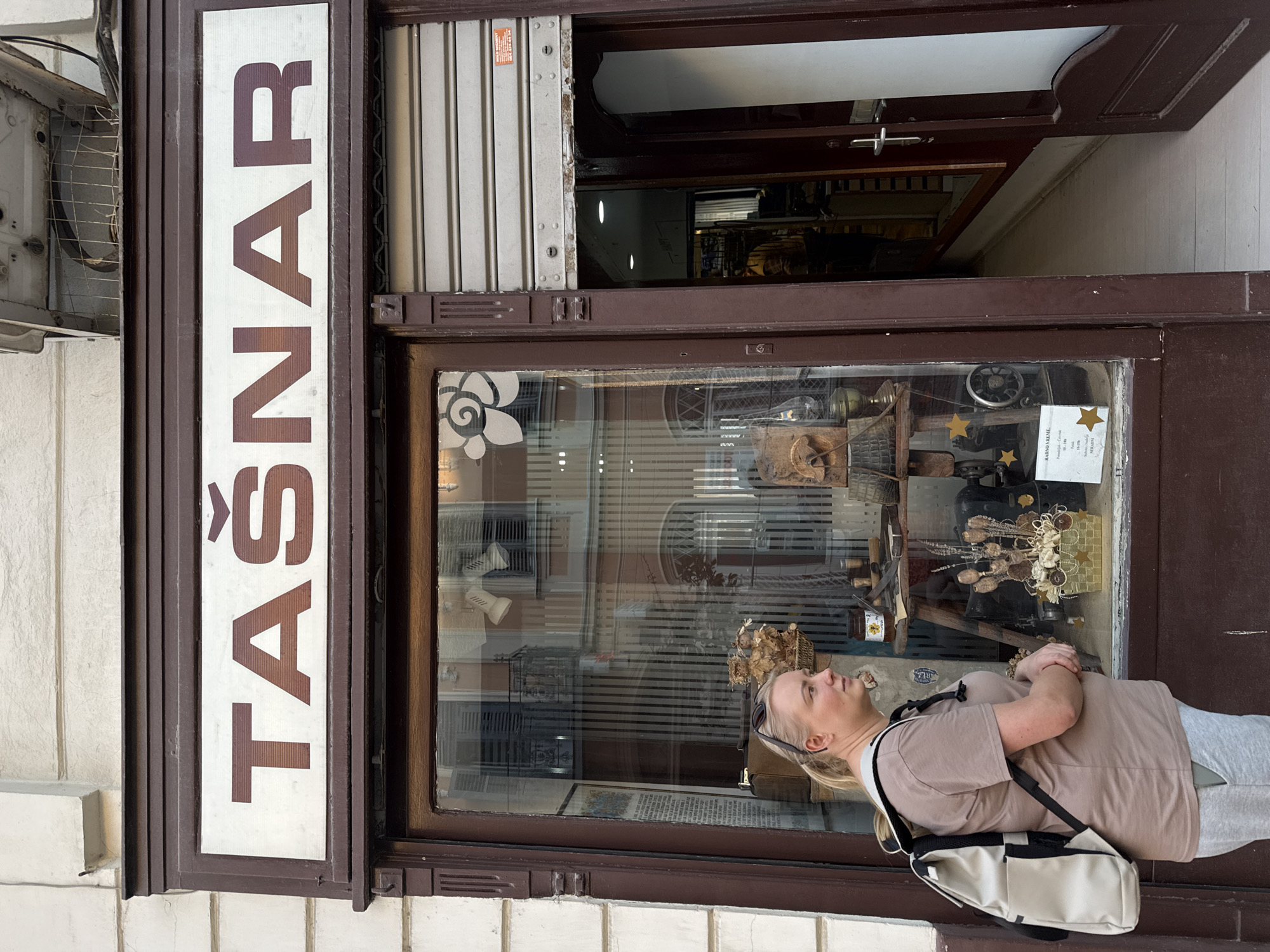 A slightly rotated outdoor shot depicts a blonde woman standing with her arms crossed in front of a shop window labeled "TAŠNAR" vertically on the left side of the frame. The shop window displays various items, including vintage objects, sewing machines, and decorative items. A partially open door is visible on the upper right, revealing the shop's interior. The building's facade is light-colored, and an air conditioning unit is mounted on the wall to the left of the sign. The woman is wearing a beige t-shirt, grey sweatpants, and a white backpack. The location is Unknown, Unknown.