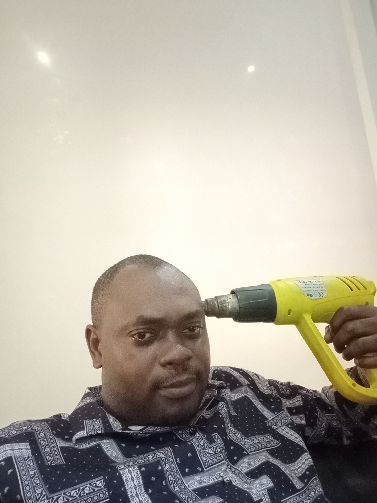 A man is holding a yellow heat gun to his forehead. The man is wearing a dark blue patterned shirt. He is in a room with a light-colored wall. There is no way to verify the location is Gwarinpa, Nigeria.