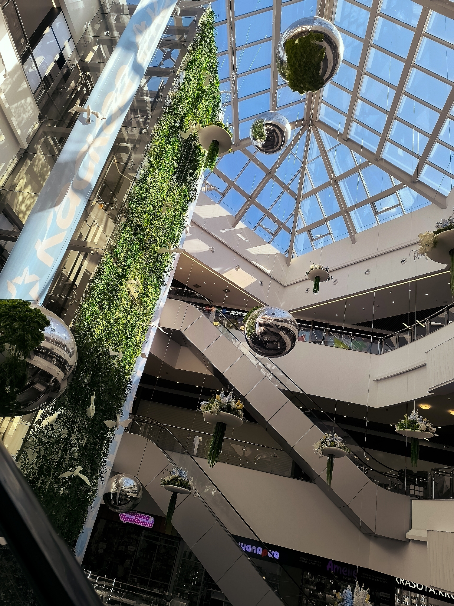 The image shows the interior of a modern shopping mall, likely in Qarağandı city administration, Kazakhstan, featuring a multi-level atrium with escalators and glass railings. A prominent vertical garden wall covered in lush green foliage runs along one side, adorned with several white bird sculptures. Large, reflective spherical decorations are suspended from the ceiling by thin wires, creating interesting visual reflections of the mall's interior and the glass-paneled roof. The roof itself is a large, geometric structure composed of glass panels, allowing natural sunlight to filter into the space. On the lower levels, shop fronts are visible with illuminated signage, including one that reads "Амелия Cosmetics" and another with Cyrillic text that appears to be "Счастье Праздника". The overall atmosphere is bright and airy, indicative of a well-maintained commercial space. No individuals are clearly discernible in the frame, though the presence of escalators suggests potential shopper activity.