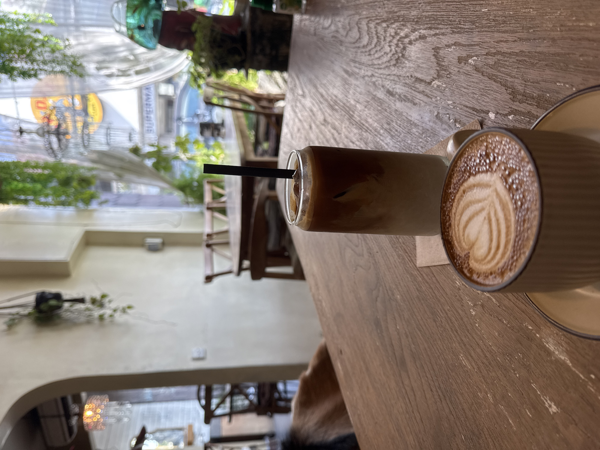 Here is a description of the image:
The image depicts a close-up of two coffee beverages on a wooden table, likely in an urban cafe setting. The scene is brightly lit, with natural light filtering in from a window or doorway visible in the background.
On the right side of the frame is a latte in a ceramic mug, with latte art in the shape of a heart adorning the surface. The mug sits on a matching saucer. To the left of the latte, there's a taller glass containing an iced coffee, identifiable by the distinct layers of coffee and milk. A black straw is inserted into the glass.
The wooden table has a textured surface, possibly meant to mimic the look of aged or reclaimed wood. The background shows a blurred street scene with greenery, suggesting an outdoor seating area or a cafe with an open view. The building across the street has hanging flower decorations.