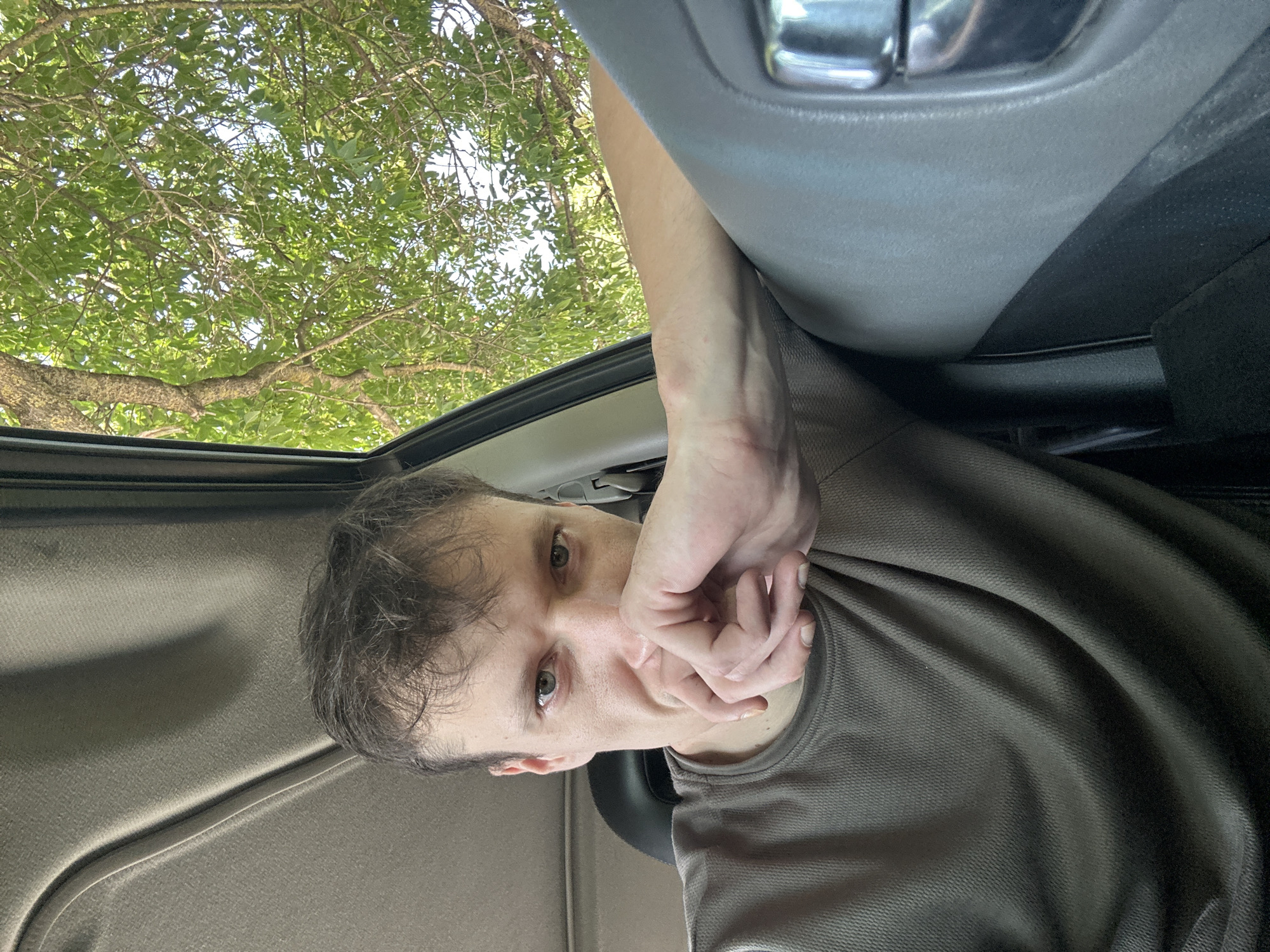 A young adult male with light skin and brown hair is captured in a close-up, selfie-style shot from within a vehicle. He is positioned in the driver's seat, leaning his left arm on the car door. His left hand is raised, with his fingers covering his mouth and nose, obscuring the lower half of his face. His green eyes are visible and directed towards the camera. He is wearing a textured, olive-green crew neck t-shirt. The interior of the car, including the ceiling lining and a portion of the dashboard, is visible. Through the open car window, a leafy green tree is seen against a bright sky. The environment appears to be outdoors, possibly in a rural or suburban setting, with the lush foliage suggesting a warm season. There is no indication of the specific location within Makiv, Ukraine.