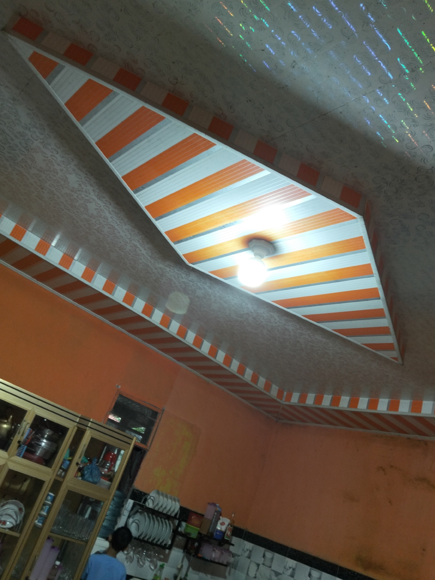 This media file captures an interior shot of a room, likely a kitchen given the presence of a cabinet filled with dishes and glassware. The ceiling is a prominent feature, decorated with a patterned texture and accented with a striking design of alternating orange and white stripes. A single light fixture hangs from the ceiling, illuminating the space. The walls are painted a vibrant orange.
In the foreground, a person wearing a blue shirt is visible from the back, facing away from the camera and towards the cabinet. The cabinet itself is made of wood and glass, showcasing stacks of plates and various glasses. The overall impression is of a functional living space, possibly in a home.
The location is specified as Payagaboh, Indonesia. The lighting suggests it is daytime, though the interior setting makes it difficult to determine the exact time of day or weather conditions outside. There is no visible text within the image. The scene appears to be a candid snapshot of domestic life.