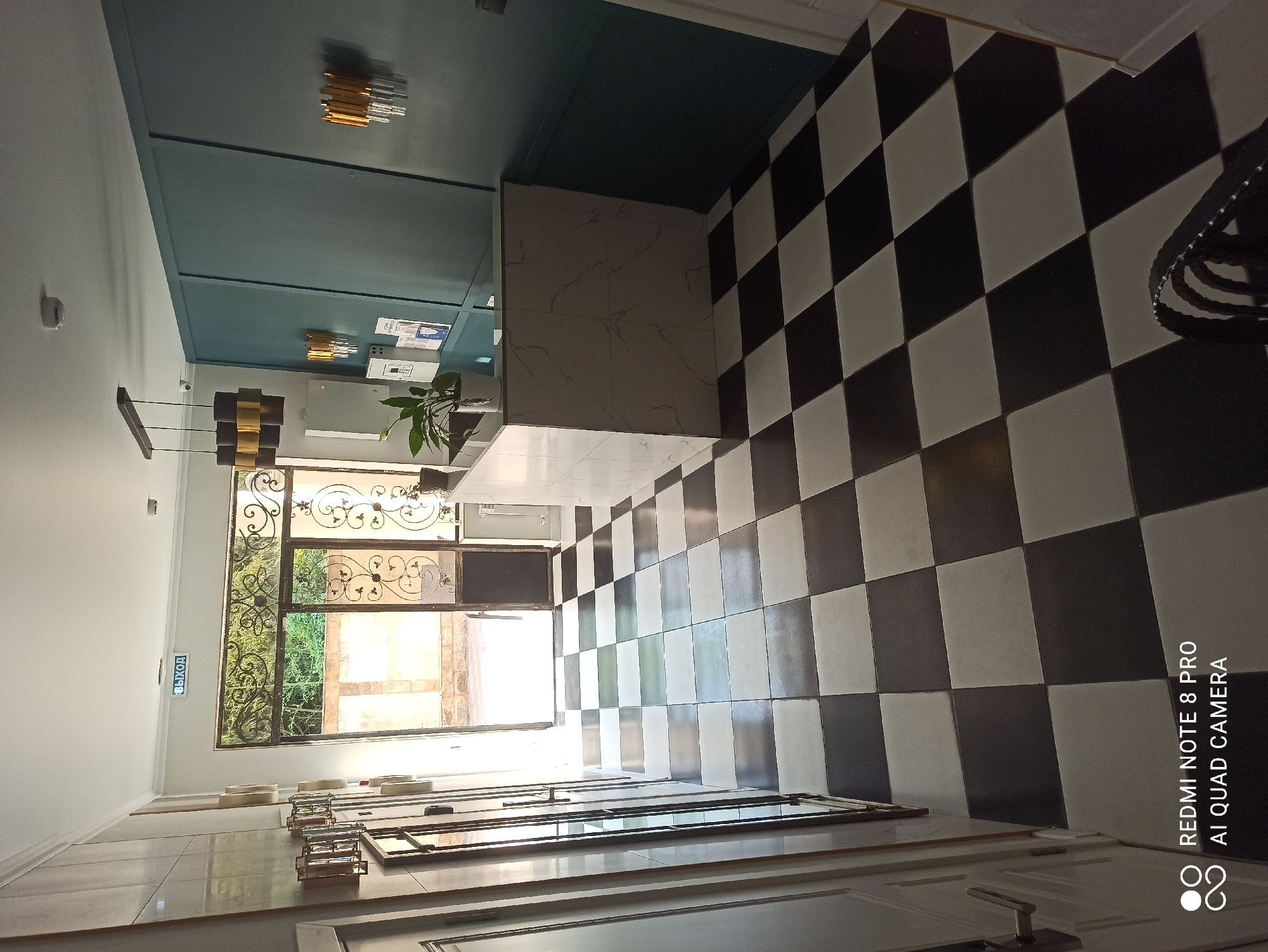 The image shows the interior of a reception area. The floor is tiled with a black and white checkered pattern. To the left, there is a white door with a handle. Further to the left, there is a wall with decorative elements and several sconces. In the center of the image, there is a reception desk with a white marbled front. Behind the desk, a wall is paneled in a dark teal color, with two gold and clear glass sconces mounted on it. A pendant light fixture with three cylindrical shades hangs from the ceiling above the desk. A green potted plant sits on the desk. To the right of the reception desk, there is a doorway with decorative metalwork leading outside to a courtyard with trees and a stone wall. An illuminated sign reading "ВЫХОД" (EXIT) is visible above the doorway. The scene appears to be a hotel or a similar establishment.