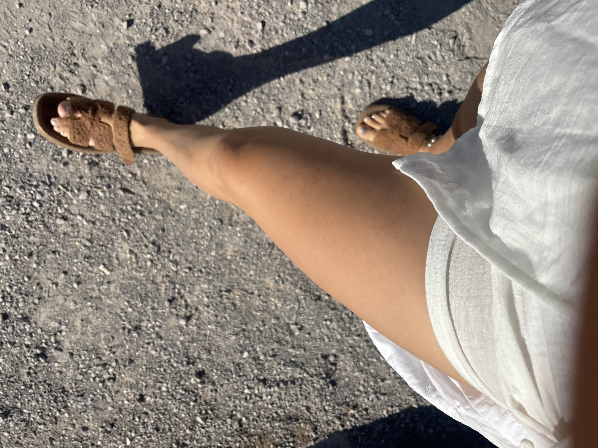 A person's legs and feet are visible in the image, standing on a gravel path. The person is wearing brown furry slides and a white shirt or dress. The sun is casting a shadow of the person onto the path, indicating it is daytime. The surroundings appear to be an outdoor area, likely in Budva, Montenegro, given the gravelly texture of the ground which is common in certain parts of the city and its outskirts.