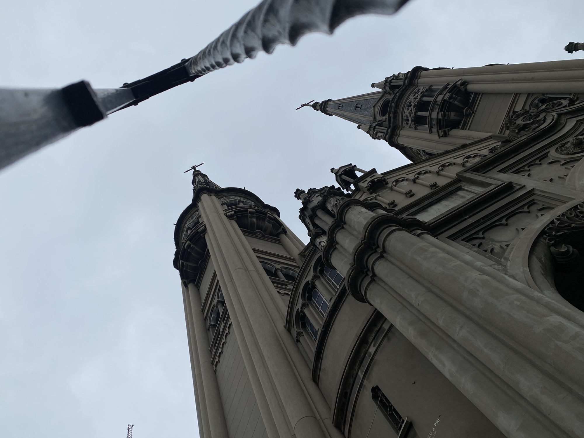 A low-angle shot captures the Metropolitan Cathedral of Buenos Aires against a cloudy sky. Three towers of the cathedral are visible, each topped with a cross. The architecture is ornate, with detailed carvings and gothic elements. A metal cable runs diagonally across the top left corner of the frame, partially obscuring the view. The location is the city of Buenos Aires, Argentina.