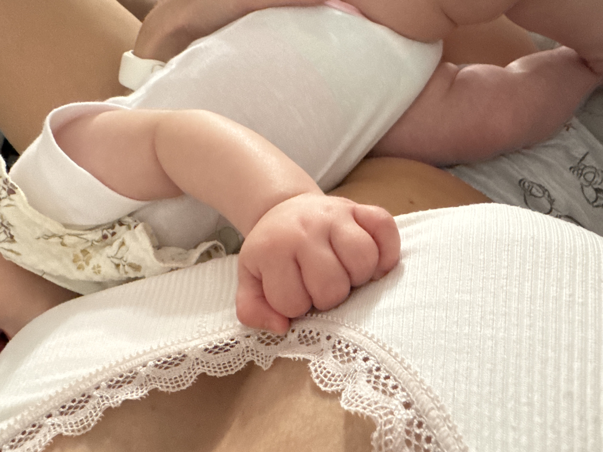 This is a close-up, low-angle, indoor shot of a mother holding her baby. The mother is wearing a white ribbed tank top with lace trim. The baby is wearing a white onesie with a floral pattern on the shoulder. The baby's arm and hand are visible, resting on the mother's chest. The baby's legs are also visible. The background is blurred, but appears to be a bed or couch with a patterned sheet. There is no identifiable location within the city of Isola delle Femmine, Italy visible in this image.