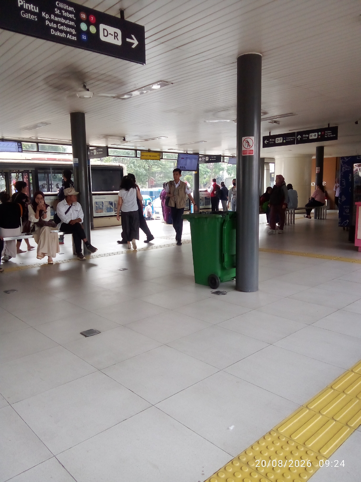 The image captures a scene at a bus stop in East Jakarta, Indonesia. The bus stop has a covered waiting area with a white tiled floor and a white ceiling. Several people are visible, some sitting on benches, others standing or walking. A large green trash bin with wheels is prominently placed near a grey pillar. Overhead signs indicate bus routes and destinations, with "Pintu D-R" and "Pintu K-R" being legible. A bus is partially visible in the background. A tactile paving strip in yellow is present on the floor, indicating a safe pathway for visually impaired individuals. The timestamp "20/08/2026 09:24" is overlaid on the bottom right of the image.