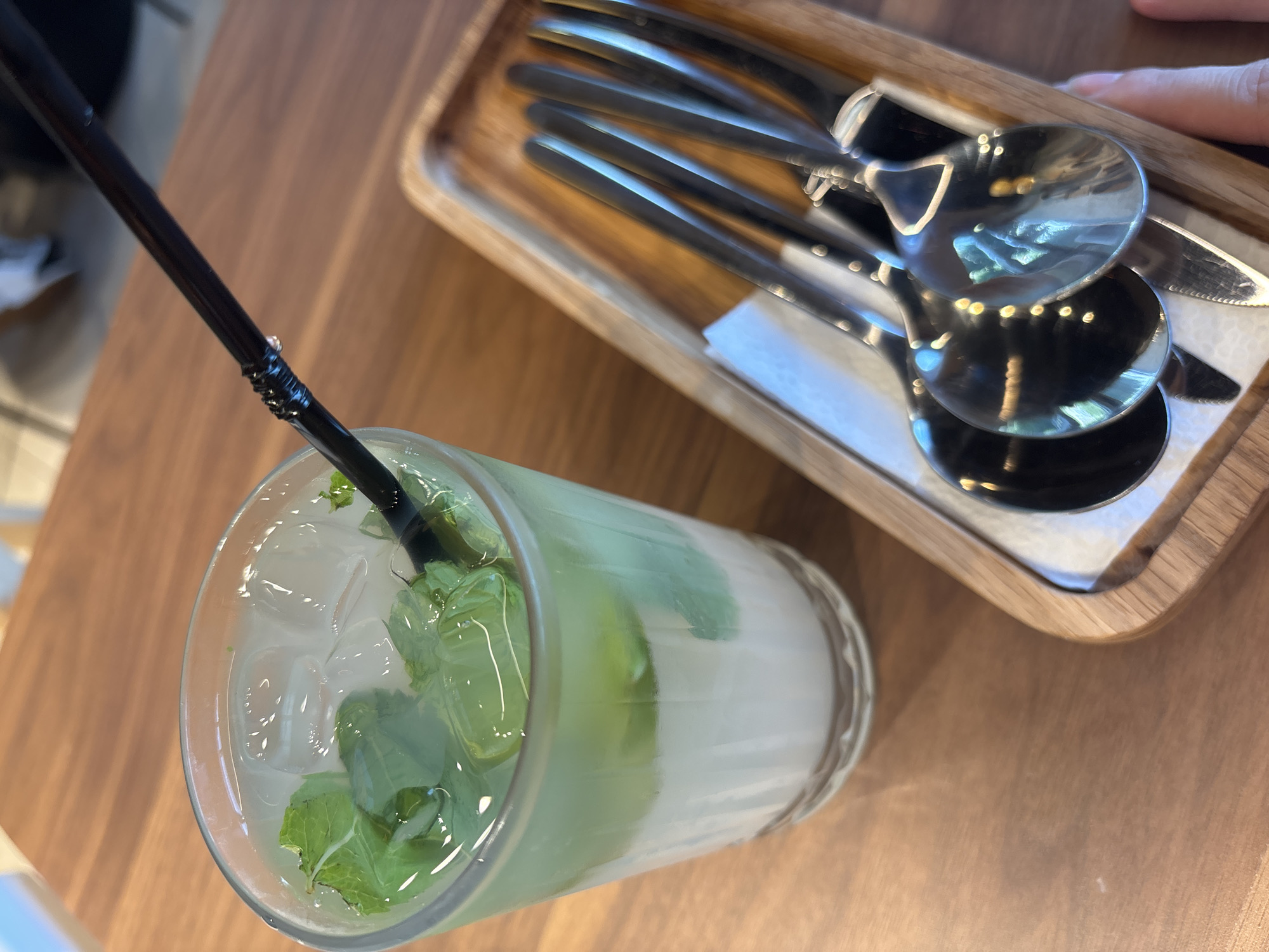A glass of mojito with mint and ice sits on a wooden table. Next to the glass, on a wooden tray, are several spoons and knives, with a white napkin underneath them. The background is blurred, showing a light-colored surface, likely a wall or another table. The scene is indoors, in what appears to be a cafe or restaurant. The city of Naberezhnye Chelny, Russia is not depicted in this image.