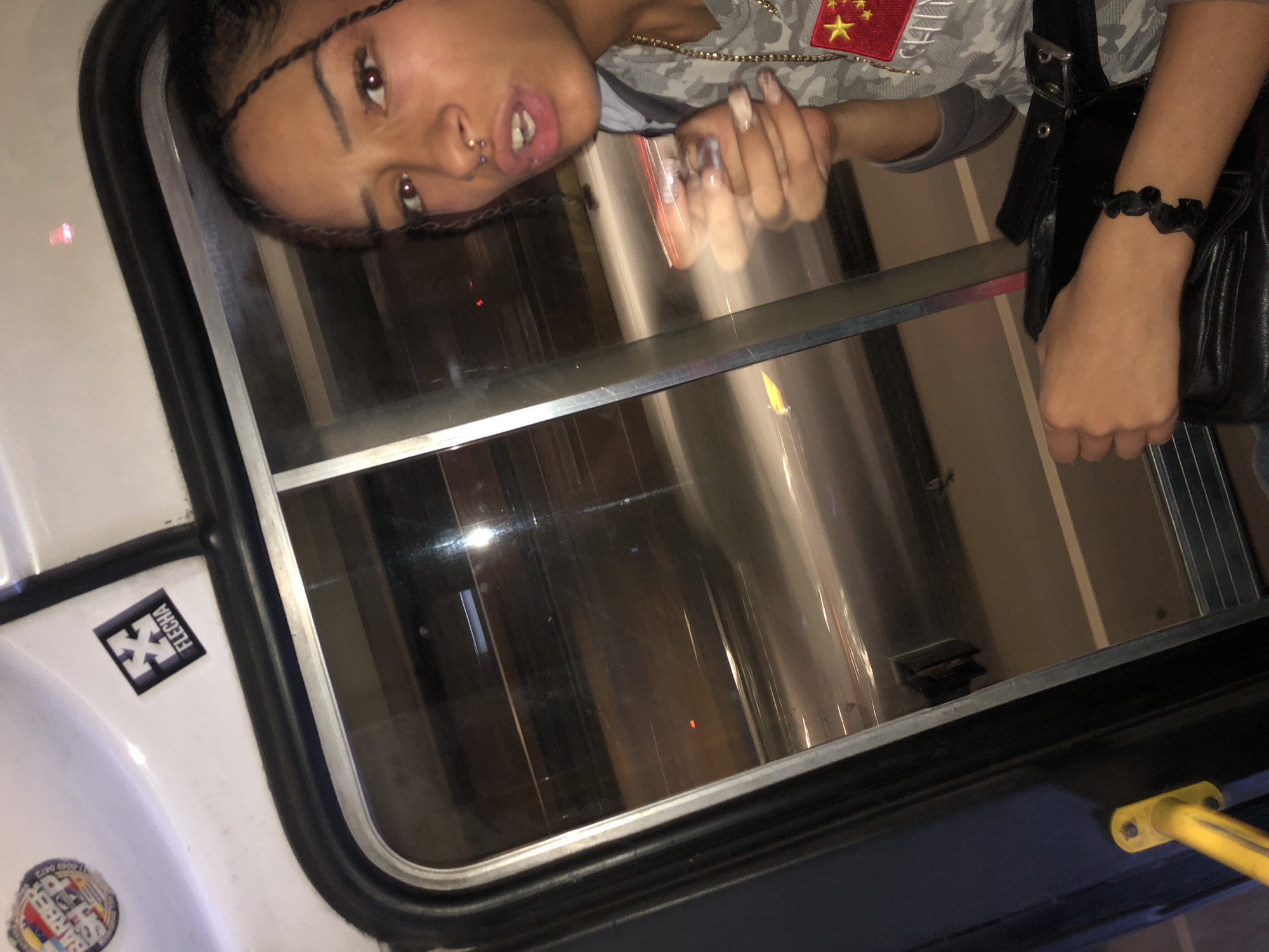A woman with braided hair and facial piercings is visible through a vehicle window. She is wearing a camouflage shirt with a Chinese flag patch. A gold chain is around her neck. She has a black purse with a black elastic bracelet on her wrist. Stickers are on the white exterior of the vehicle next to the window. A yellow pole is visible near the bottom right of the frame. It is not possible to confirm the location is Buenos Aires, Argentina.