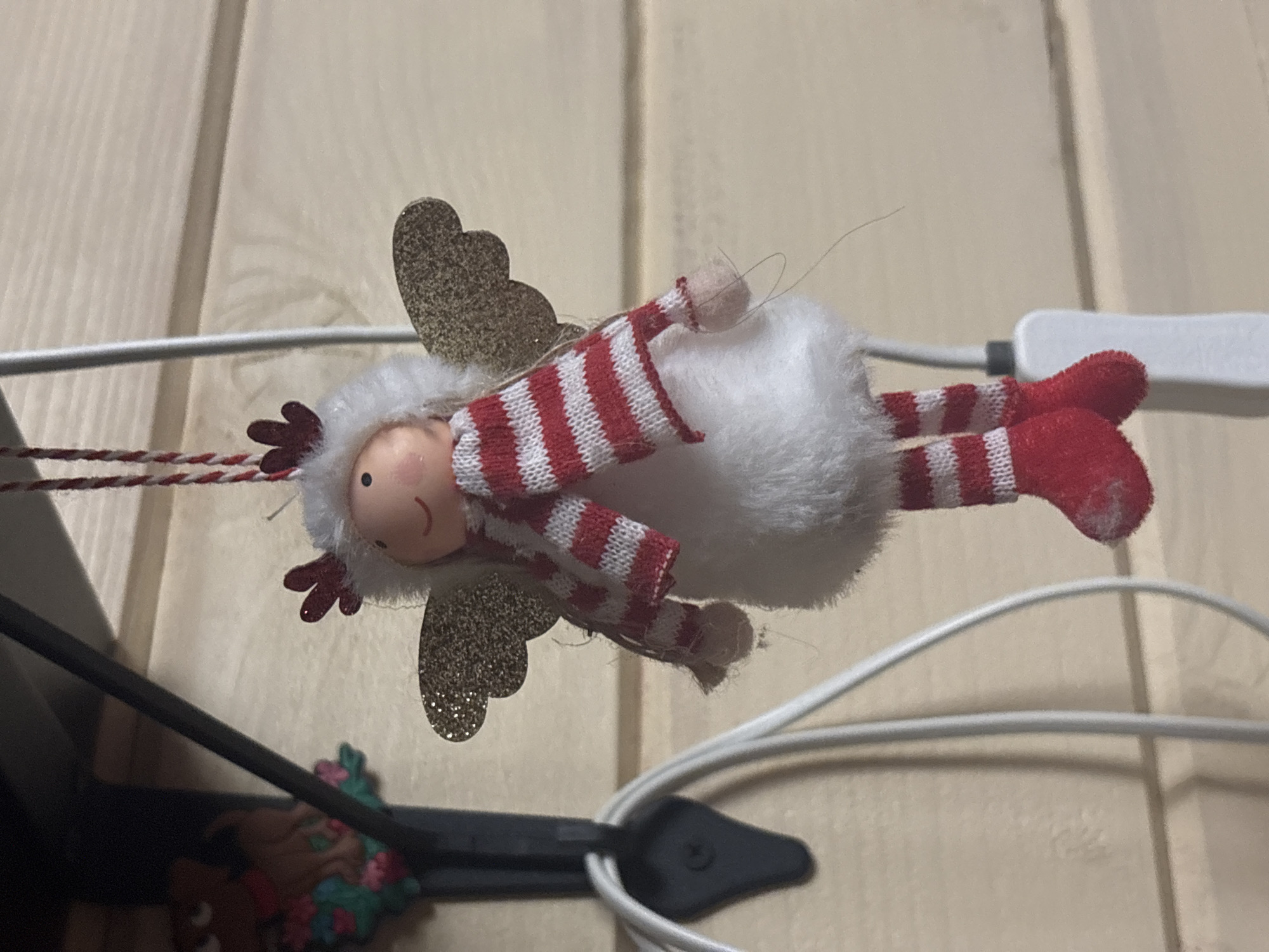 An ornament resembling a Christmas angel is hanging against a wood-paneled wall. The angel has a round face with painted eyes and a smiling mouth. It wears a white, fluffy hood with red antlers, a red and white striped scarf, a white fluffy body, and red and white striped stockings with red booties. Gold glittery wings are attached to its back. A red and white string hangs from the top, suspending the ornament. To the left of the angel, a black hook is visible, from which a white cord is hanging. In the upper left corner, partially obscured, is another ornament that appears to be a small, brown, anthropomorphic figure with red and pink floral elements. The scene is indoors, likely a home, in the city of Berezkа SNT (Povarovo), Russia.