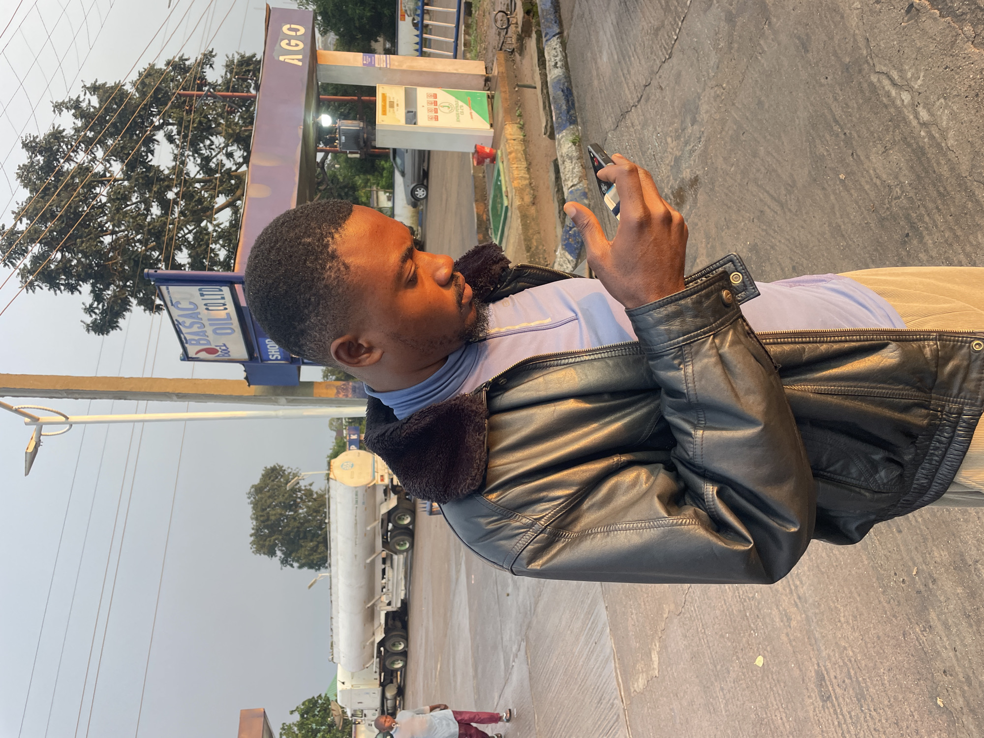 A man is standing outdoors, looking down at his mobile phone. He is wearing a black leather jacket over a light blue shirt and tan pants. The man is standing on a paved surface, with a gas station visible in the background. The gas station has pumps and signage, including "BASAC OIL CO. LTD." and "AGO". A large white tanker truck is parked in the background. The sky is overcast. The image appears to have been taken in the city of Jos, Nigeria.