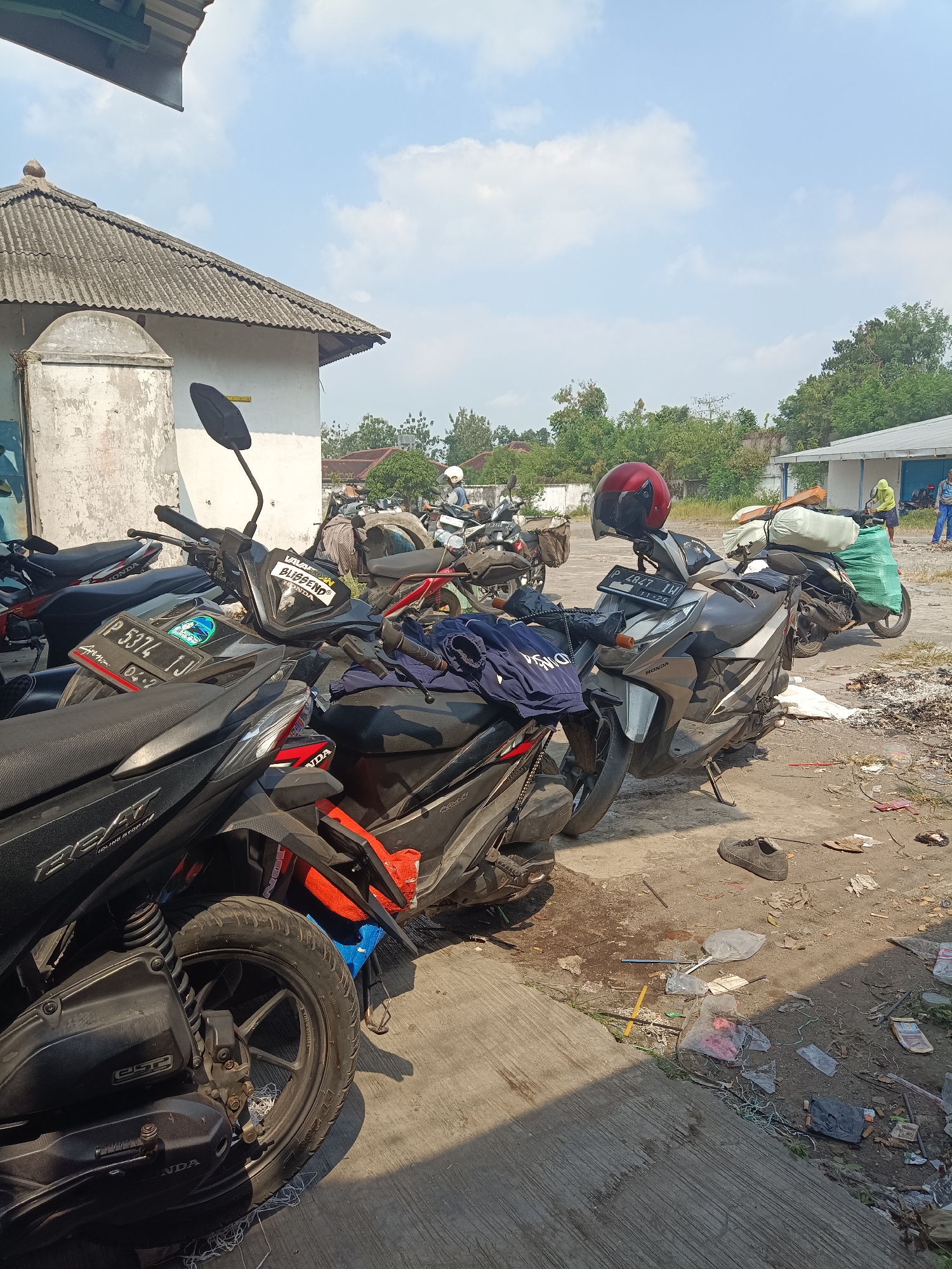 The image depicts a sun-drenched outdoor scene in Jember, Indonesia, likely a parking area. Numerous motorcycles are parked closely together, some covered with tarps or with items tied to their rear. A red helmet rests on the handlebars of a silver scooter. In the background, a white building with a corrugated roof stands, partially obscured by trees. To the right, two individuals in casual attire are visible near another building. The ground is paved and littered with debris, including plastic bags, wrappers, and discarded items, suggesting a somewhat neglected environment. The sky is partly cloudy.