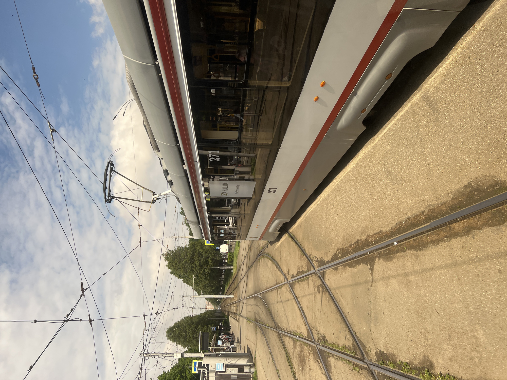 The image shows a modern tram in Krasnodar, Russia. The tram, numbered 271, is light gray with a red stripe. It is standing on a concrete tram platform, and the tracks curve in the foreground. Overhead wires crisscross above the tracks. Trees line the street in the background, and the sky is partly cloudy.